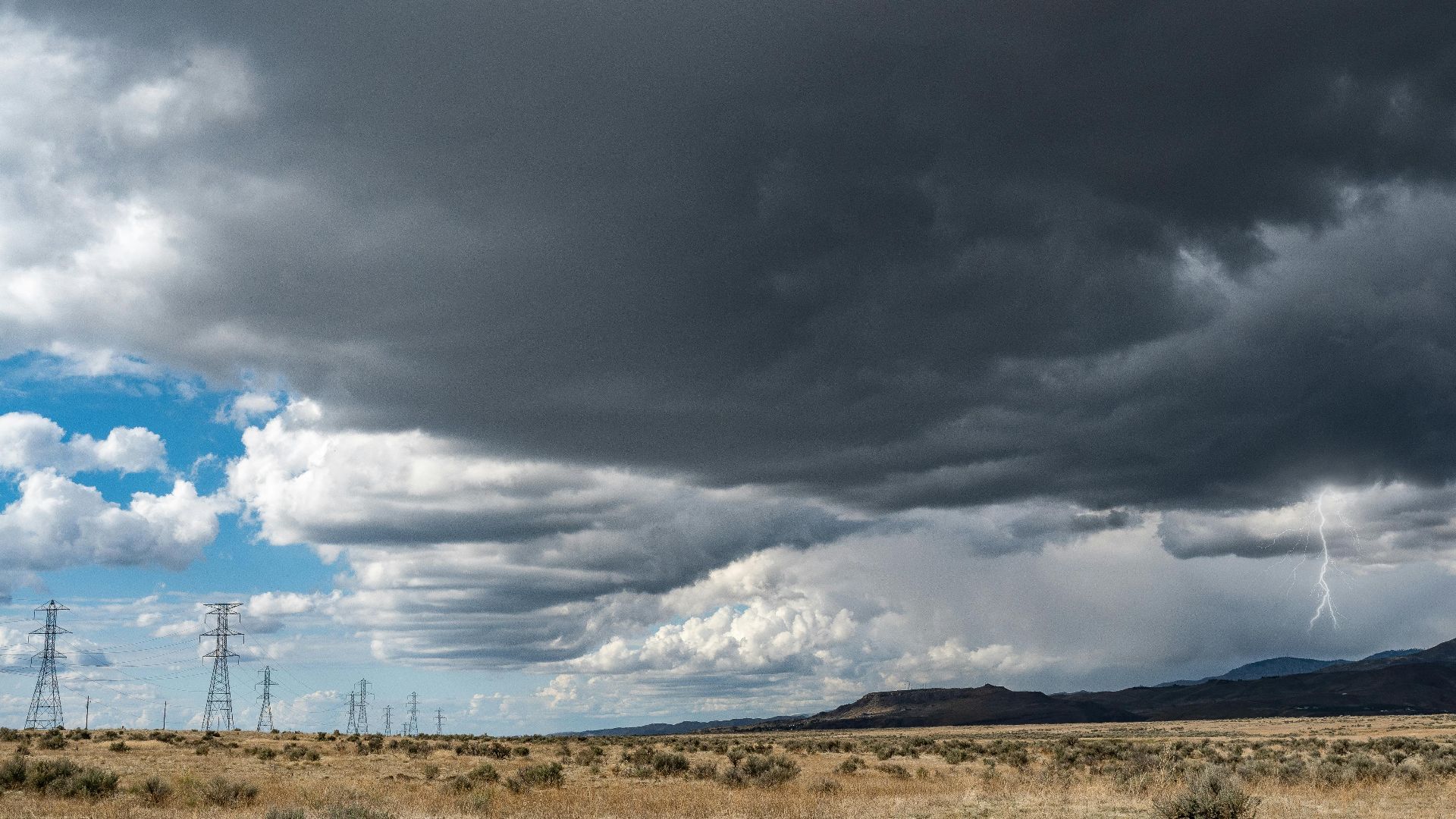 Breathtaking view of thick dark clouds on sky with lightning above uneven field with growing plants near metal towers and mountains in gloomy weather