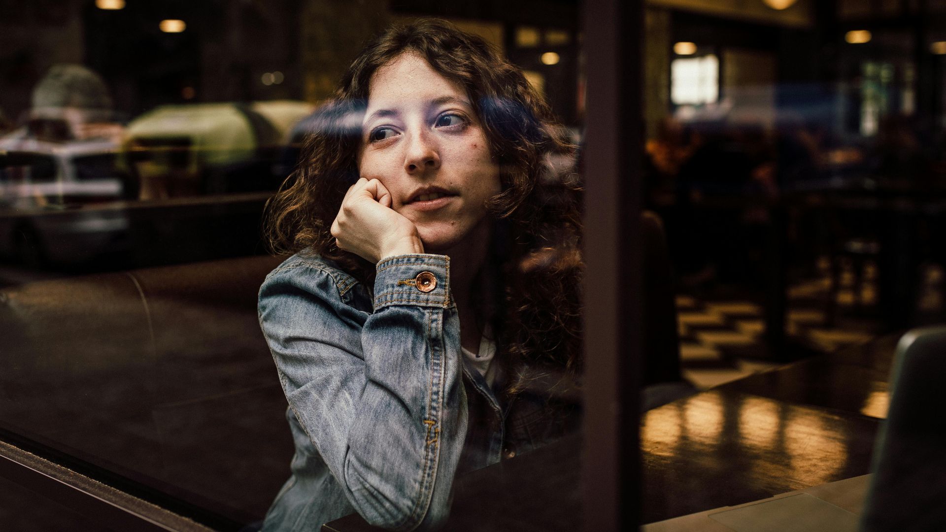 A thoughtful woman sits in a coffee shop, gazing through a window, reflecting in the glass.