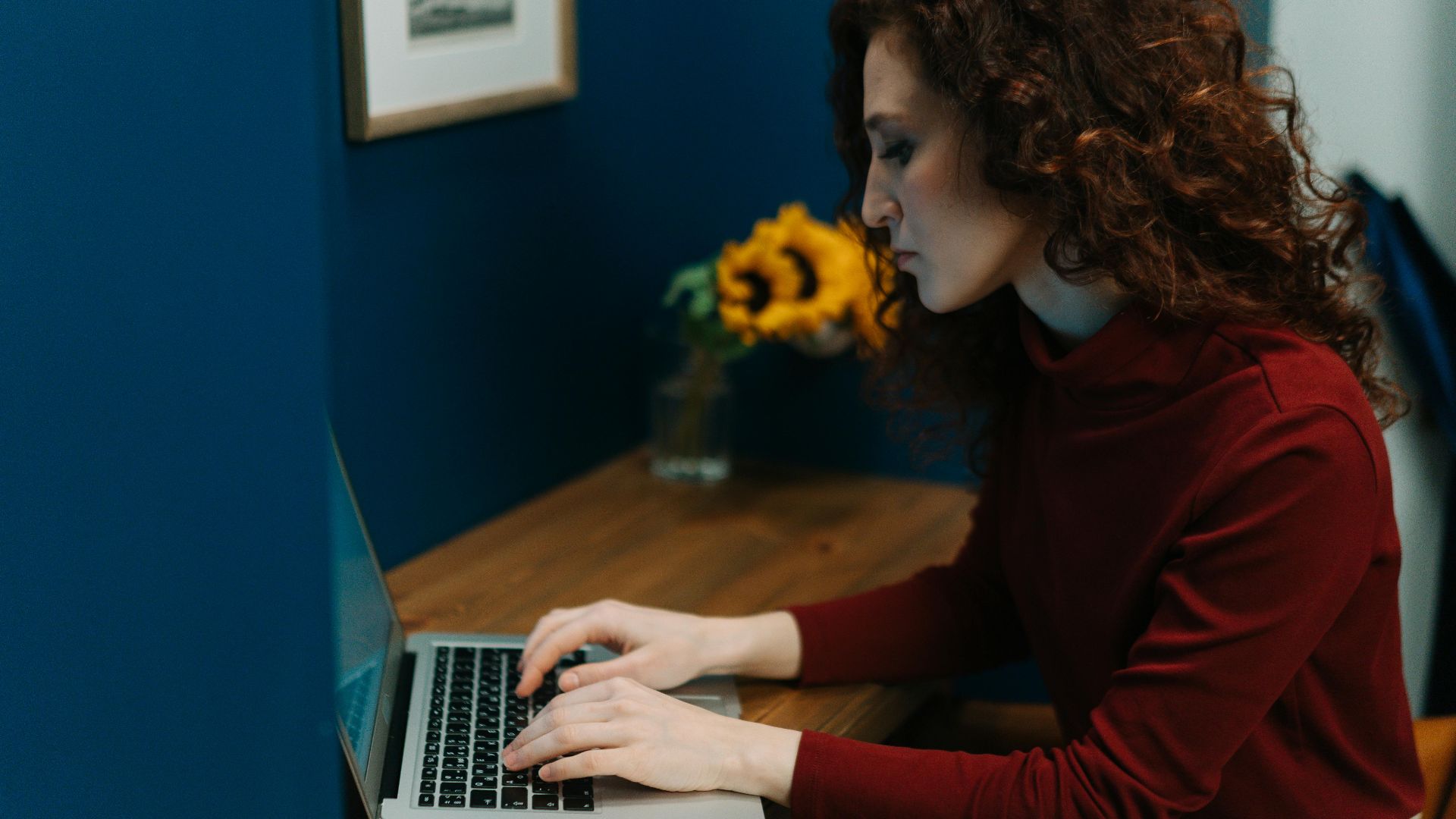 A woman working on a laptop at a desk with sunflowers, surrounded by blue decor, focusing intently.
