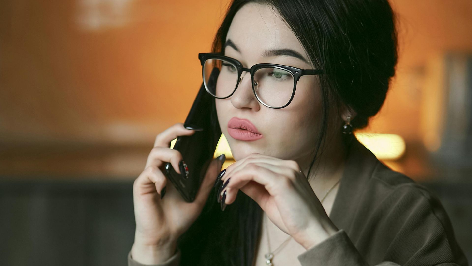A woman with eyeglasses engaged in a phone call indoors, focused and thoughtful.