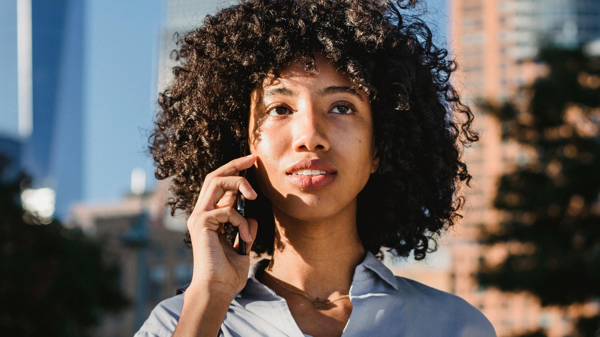 Confident young woman with curly hair talks on phone while holding coffee cup in urban setting.