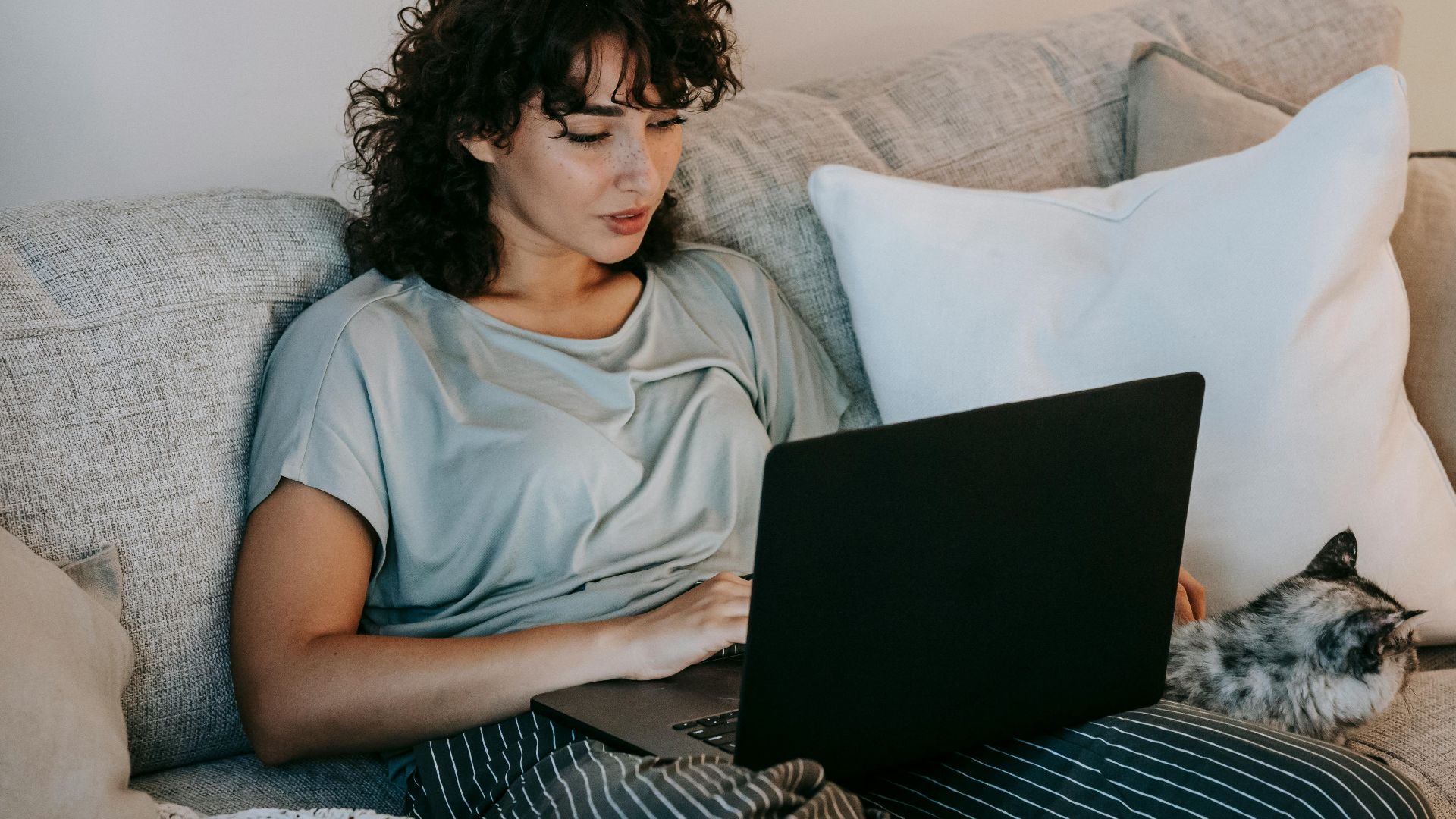 A young woman with curly hair relaxes on a cozy sofa, working on her laptop with a cat beside her.