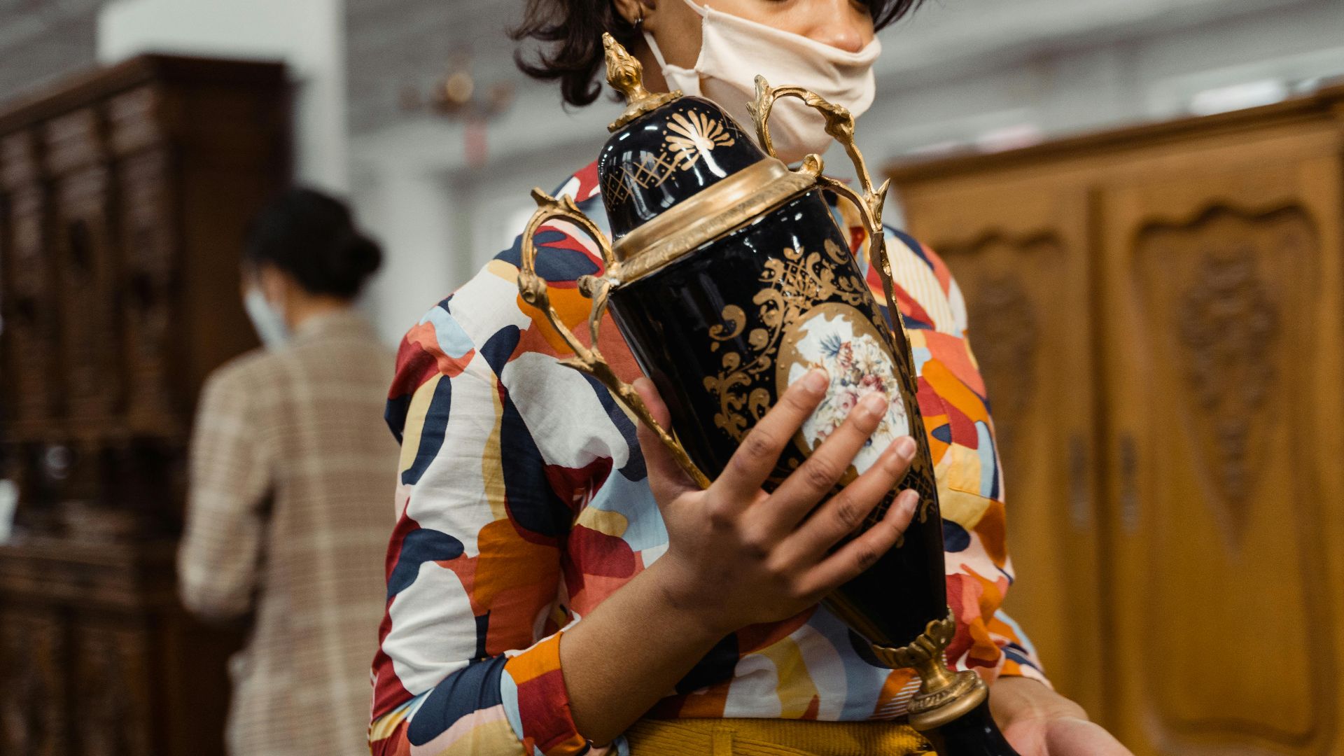 Woman wearing colorful shirt examines ornate vase in a vintage shop.