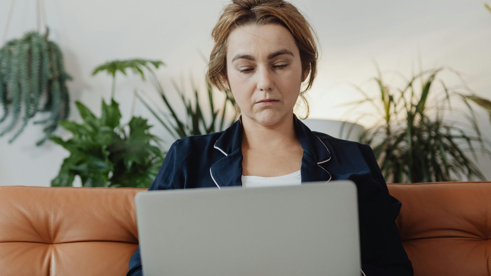 Woman in pajamas working on a laptop in a cozy living room surrounded by plants.