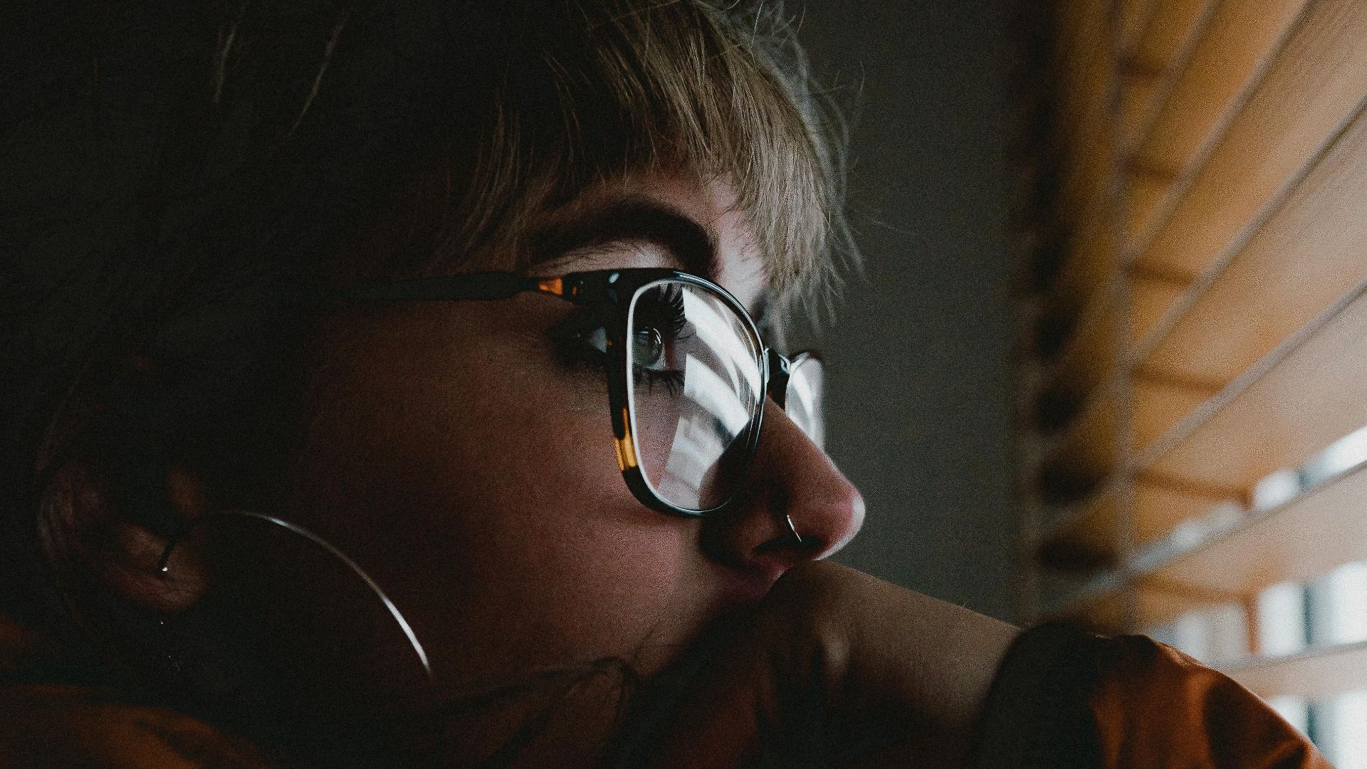 Pensive woman in eyeglasses gazing through blinds, contemplating indoors.