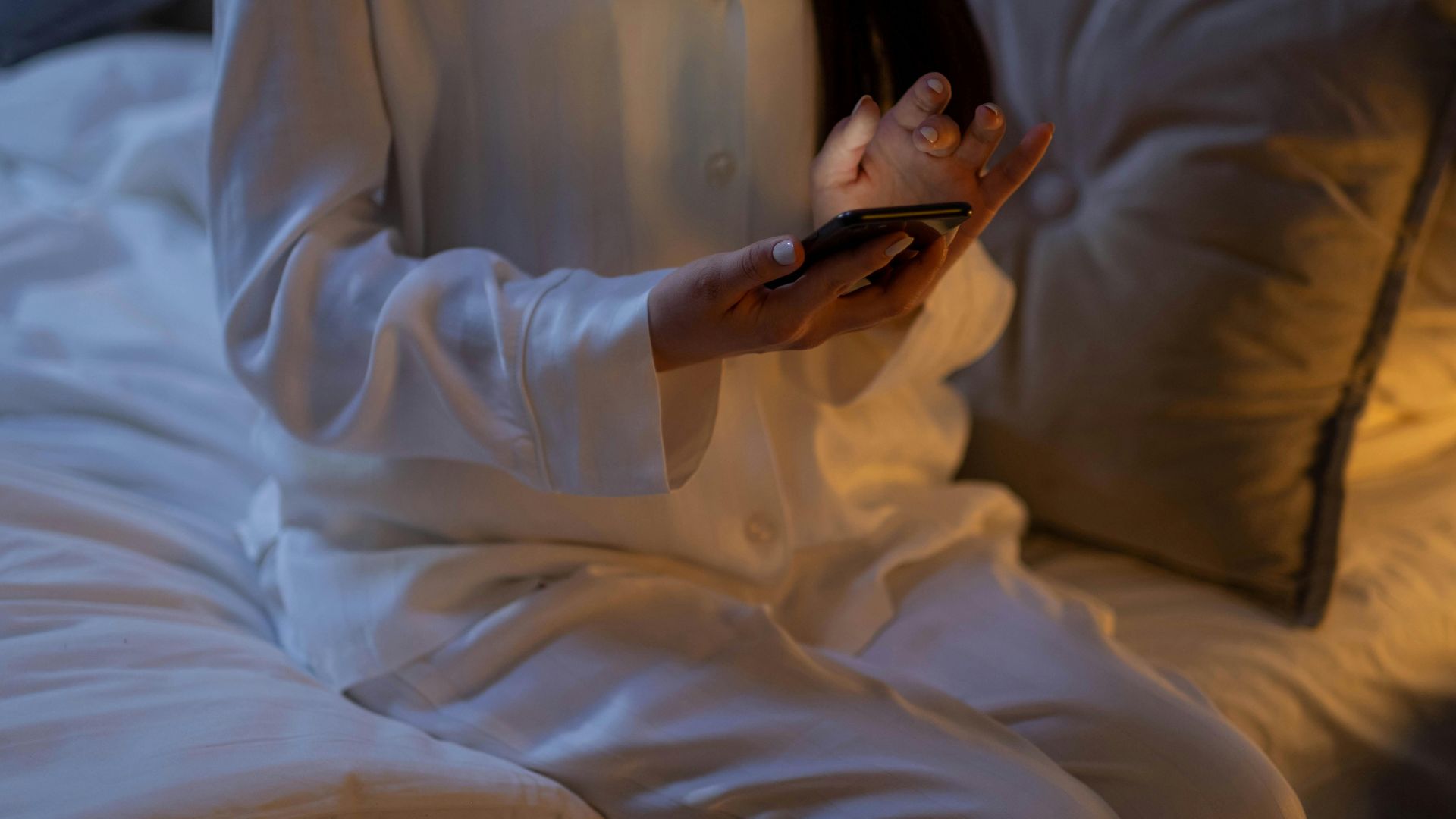 A woman in white pajamas using a smartphone while sitting on a bed at night.