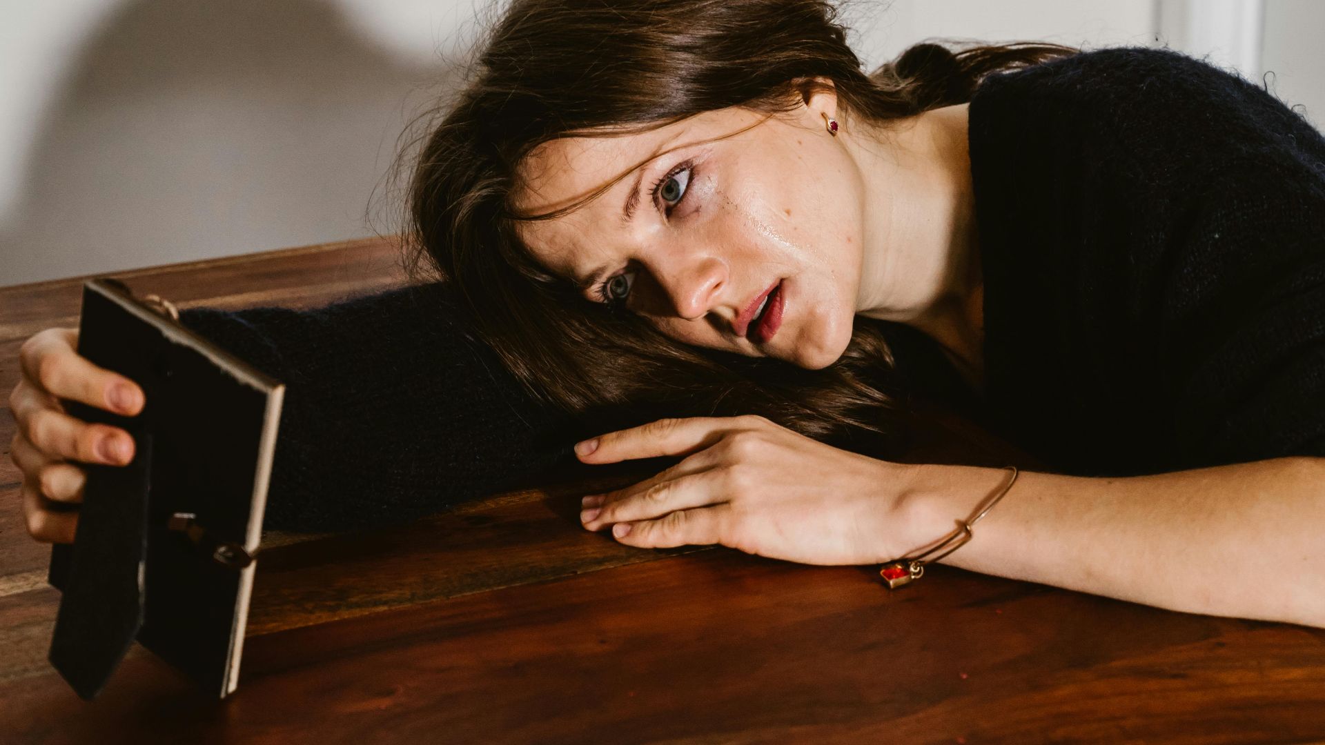 A woman in emotional distress lays on a wooden table holding a picture frame.