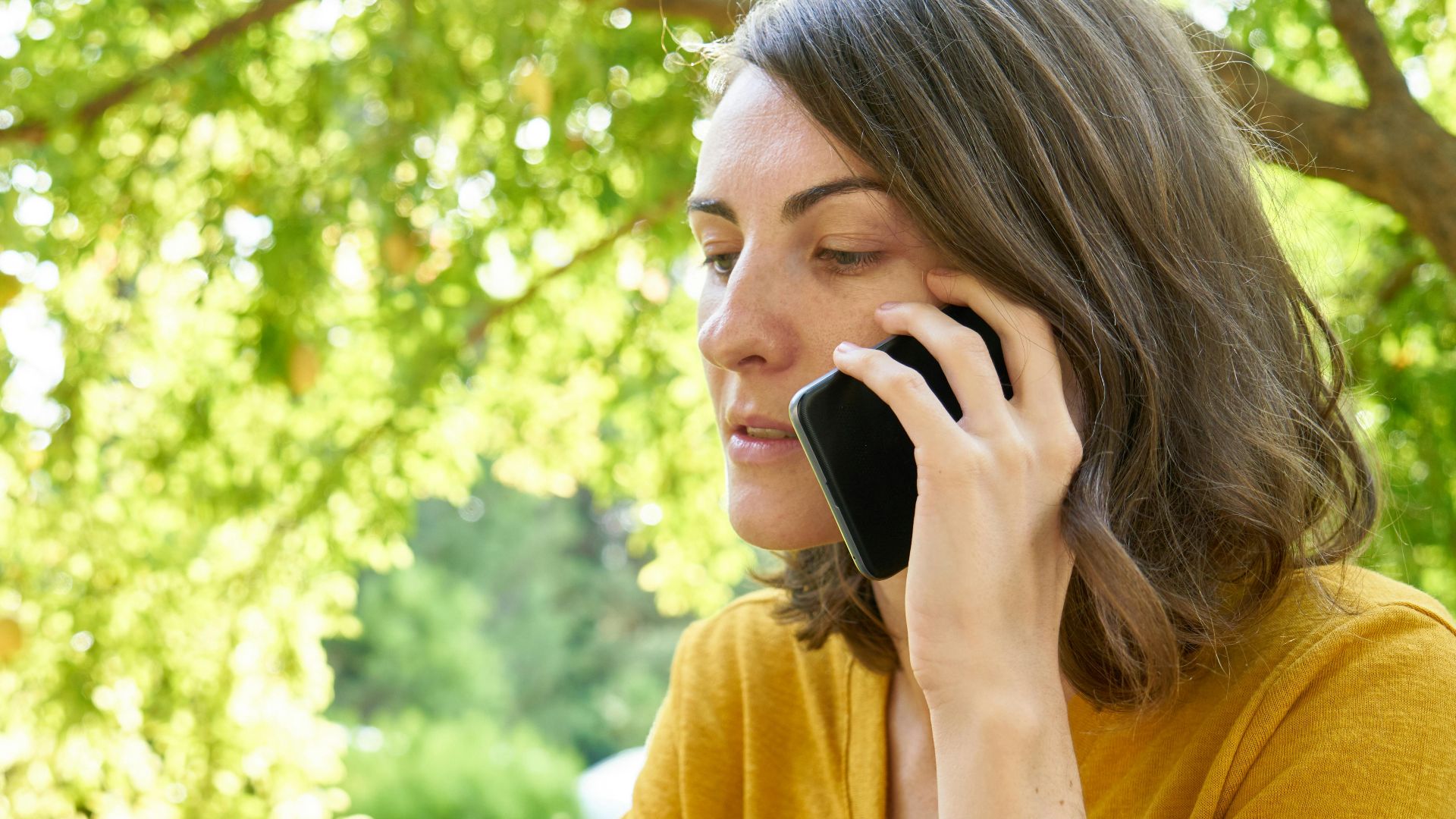 A woman in a yellow shirt makes a phone call outdoors amid lush greenery.