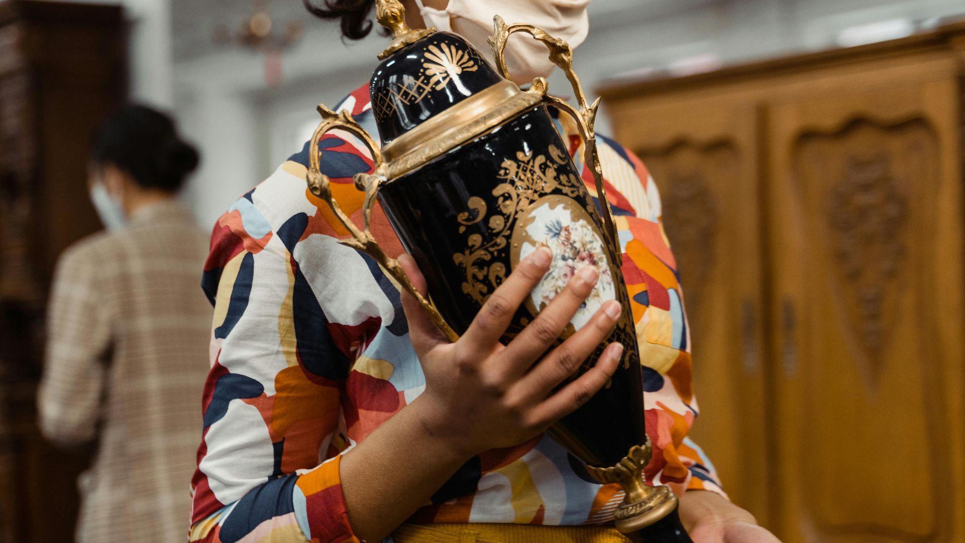 Woman wearing colorful shirt examines ornate vase in a vintage shop.
