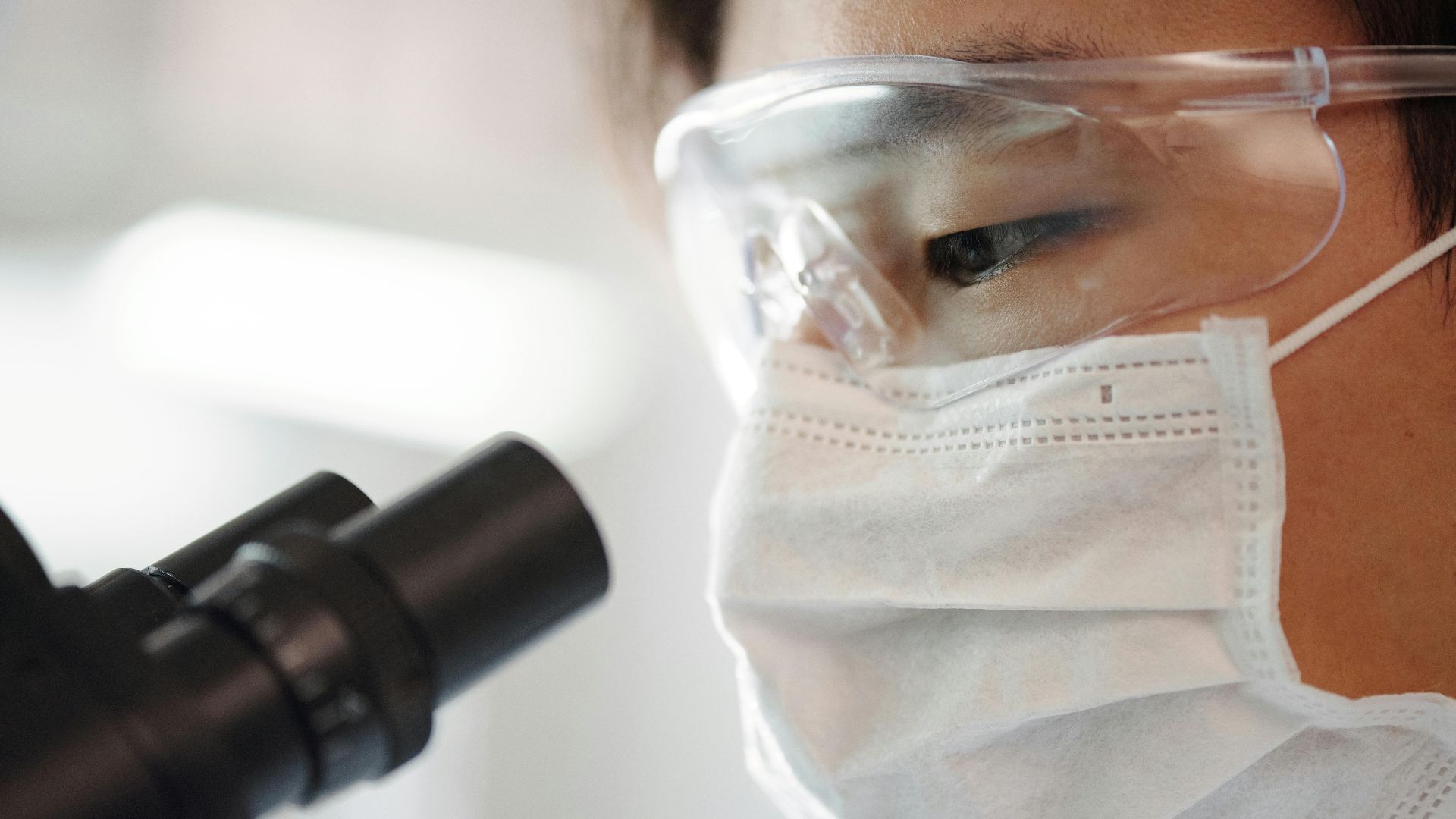Close-up of a scientist wearing safety gear using a microscope indoors.