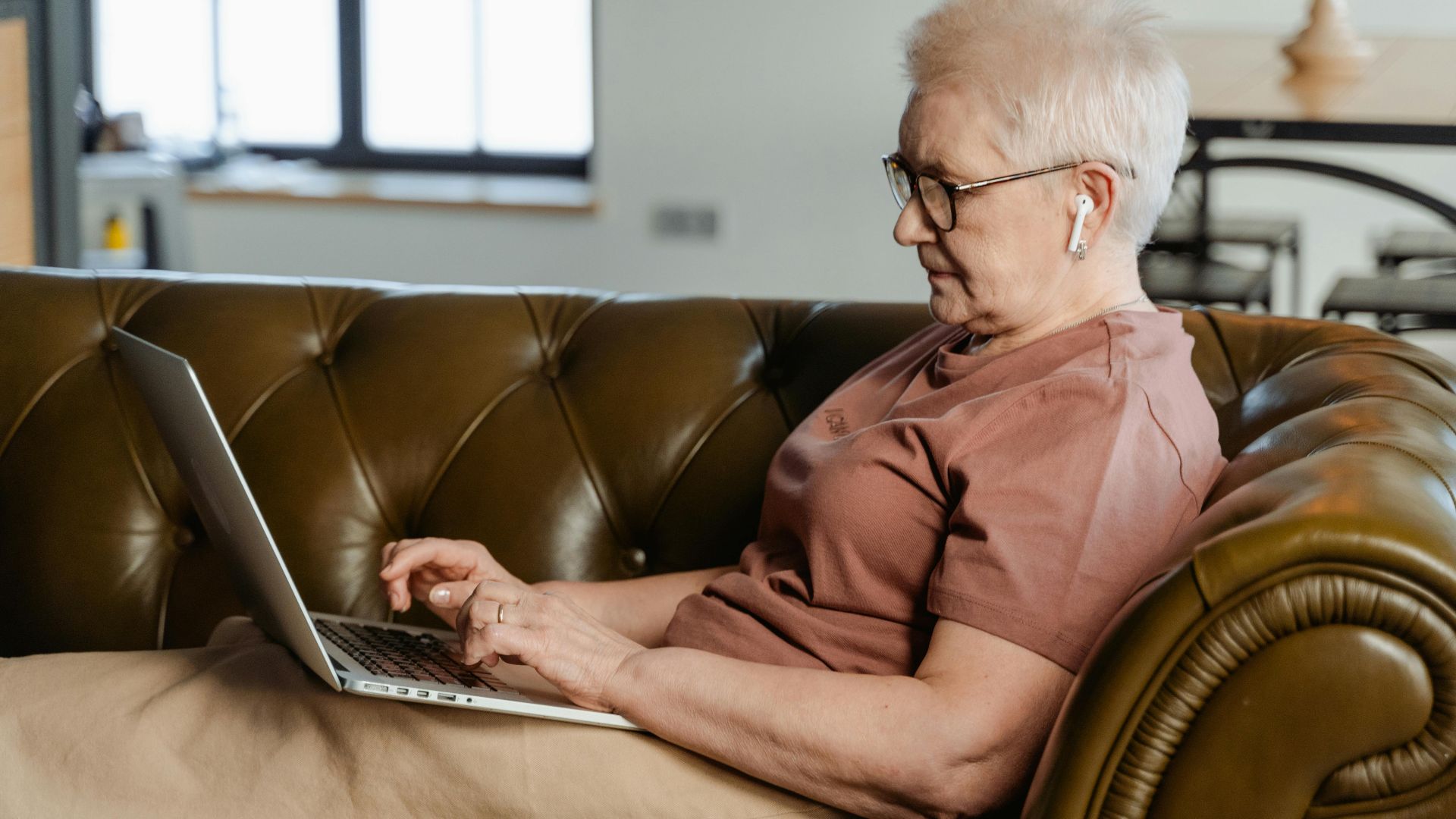 Elderly woman relaxing on a couch, using a laptop indoors during the day.