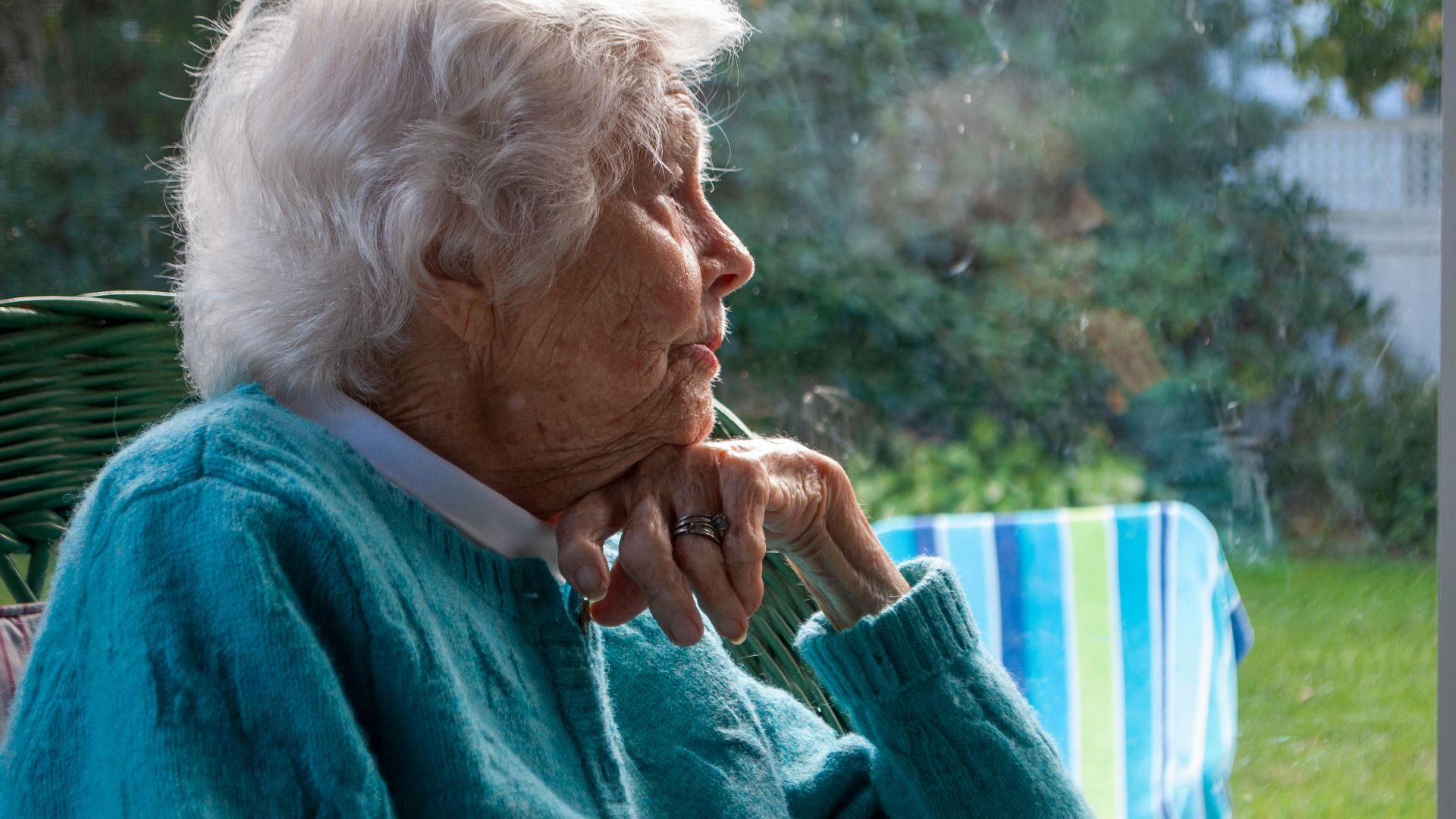 Side view of pensive senior female with white hair in warm sweater leaning on hand and looking away while resting on chair near window at home