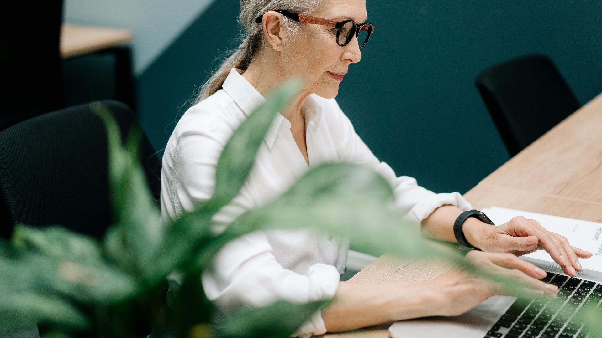 Confident senior woman with glasses working on a laptop in a modern office setting.