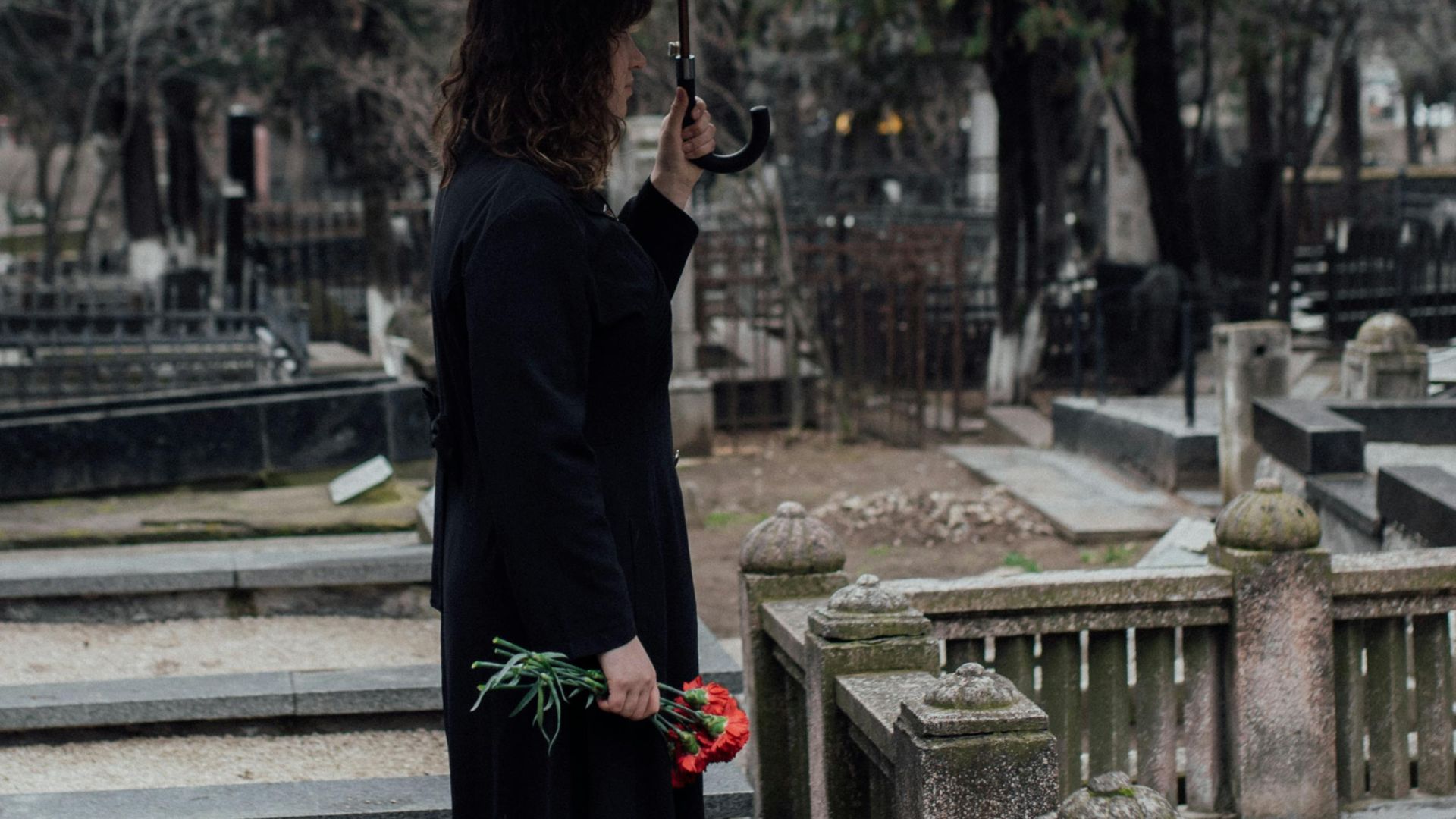 A woman stands in a cemetery with roses, holding an umbrella. Serene and reflective.
