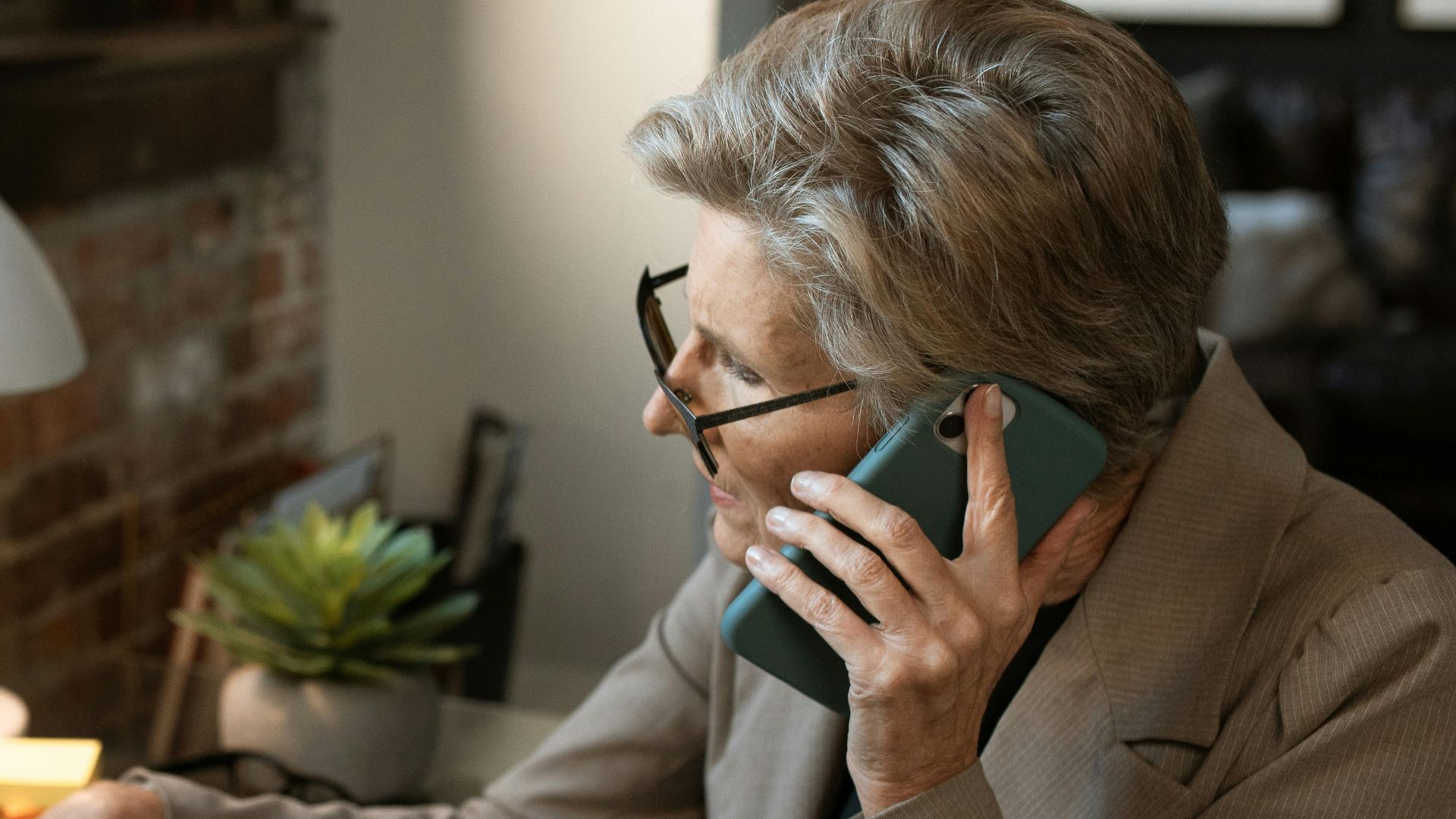 Senior woman in office working on a laptop while on the phone, demonstrating multitasking skills.