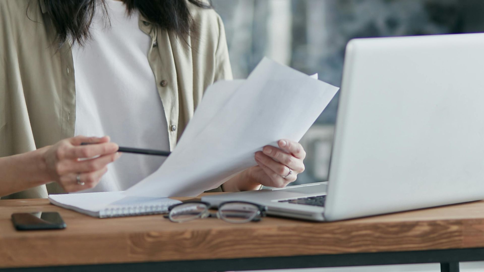 Concentrated woman reviewing documents and working on a laptop at home office setup in kitchen.