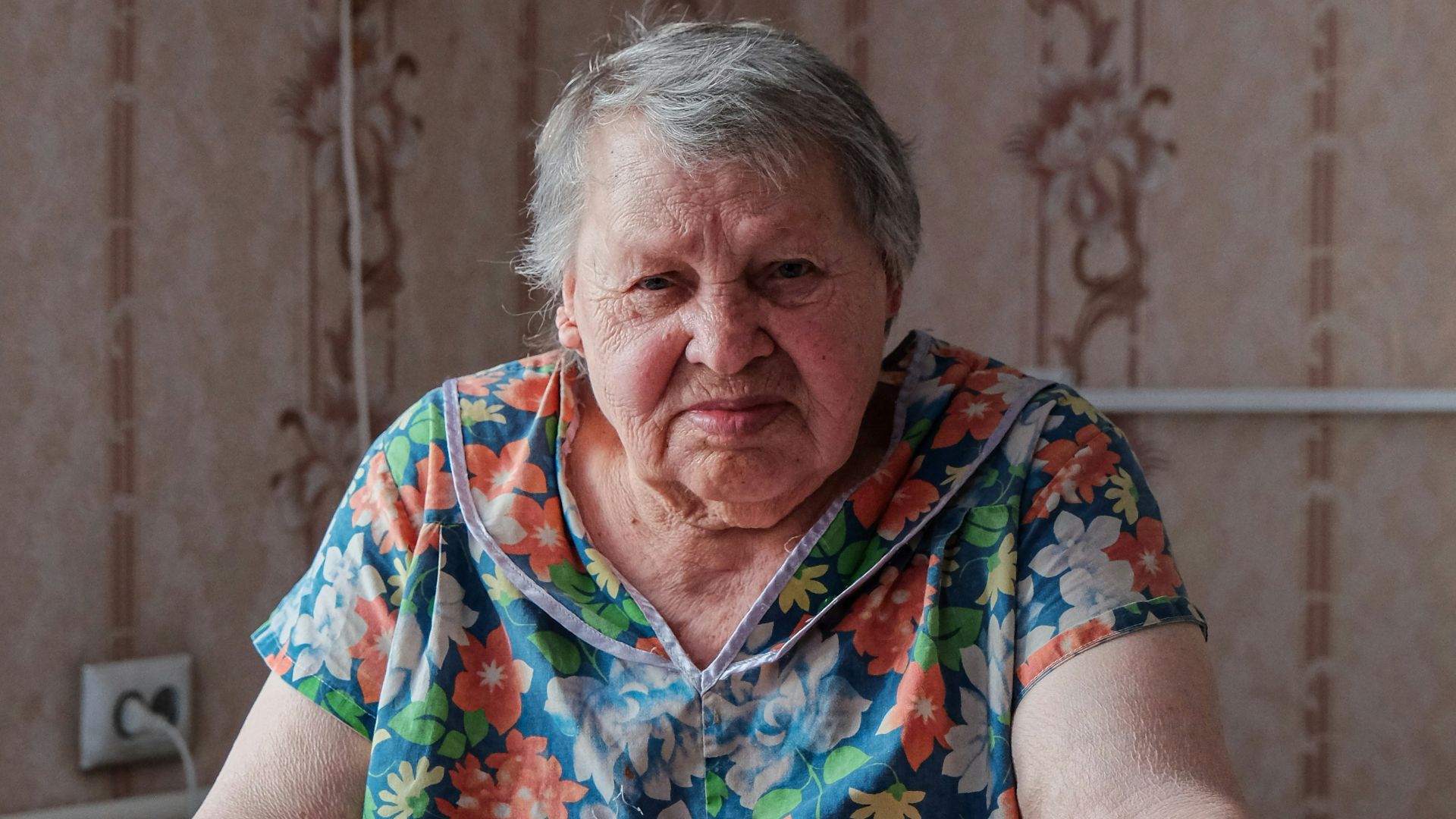 An elderly woman sits indoors, facing the camera.