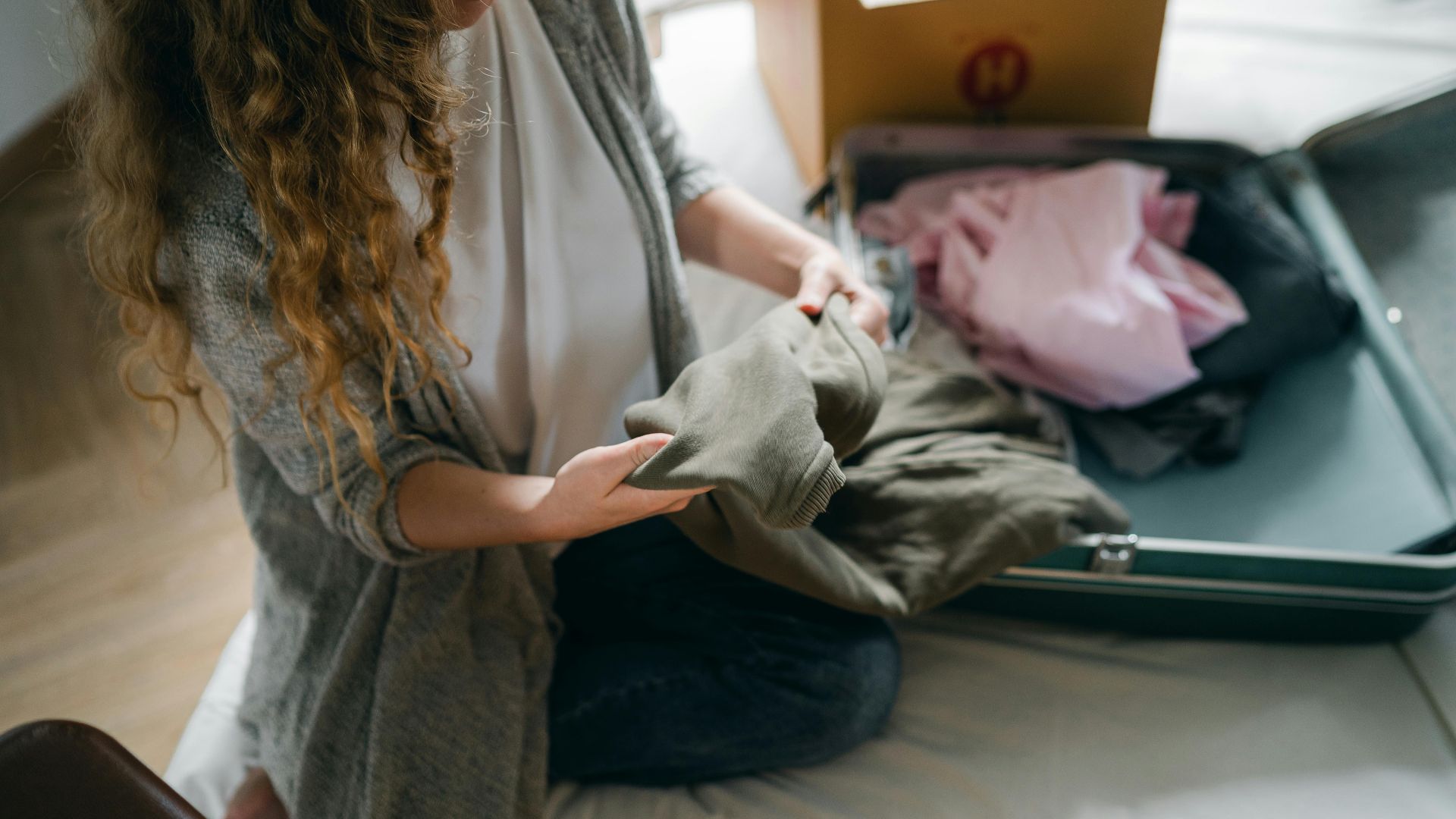 From above crop anonymous female in casual wear sitting on bed and packing luggage while preparing to move out