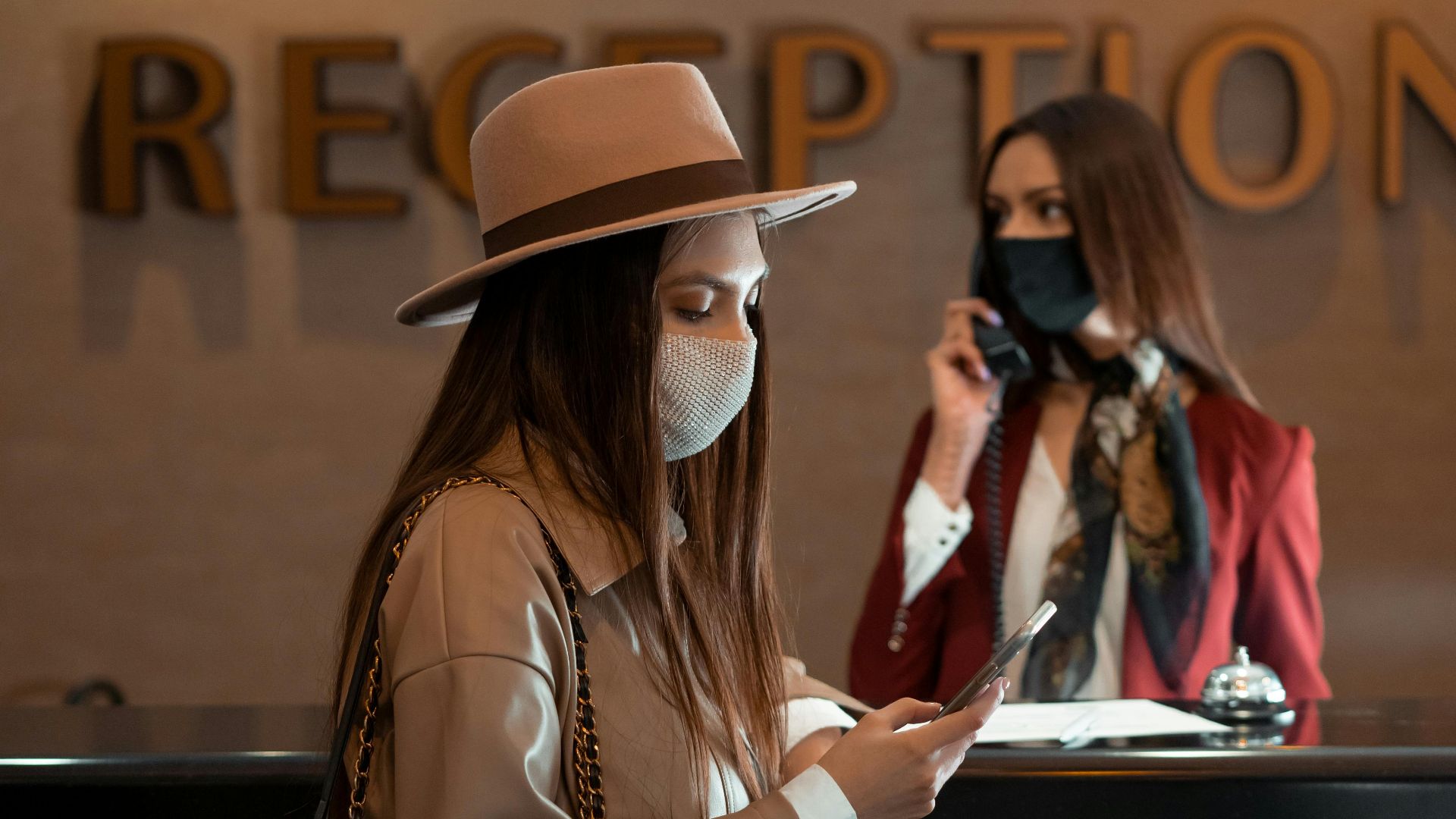 Woman in face mask at hotel reception counter checking her phone, symbolizing the new normal.