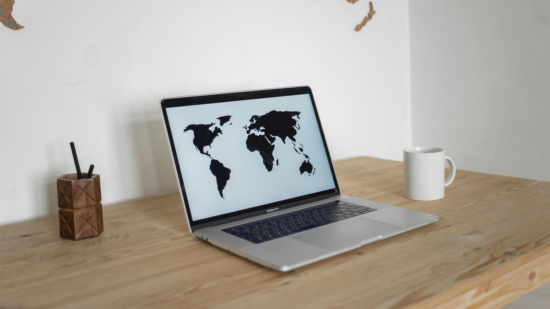A minimalist desk setup with a laptop showing a world map screen and a mug, set against a wall map backdrop.