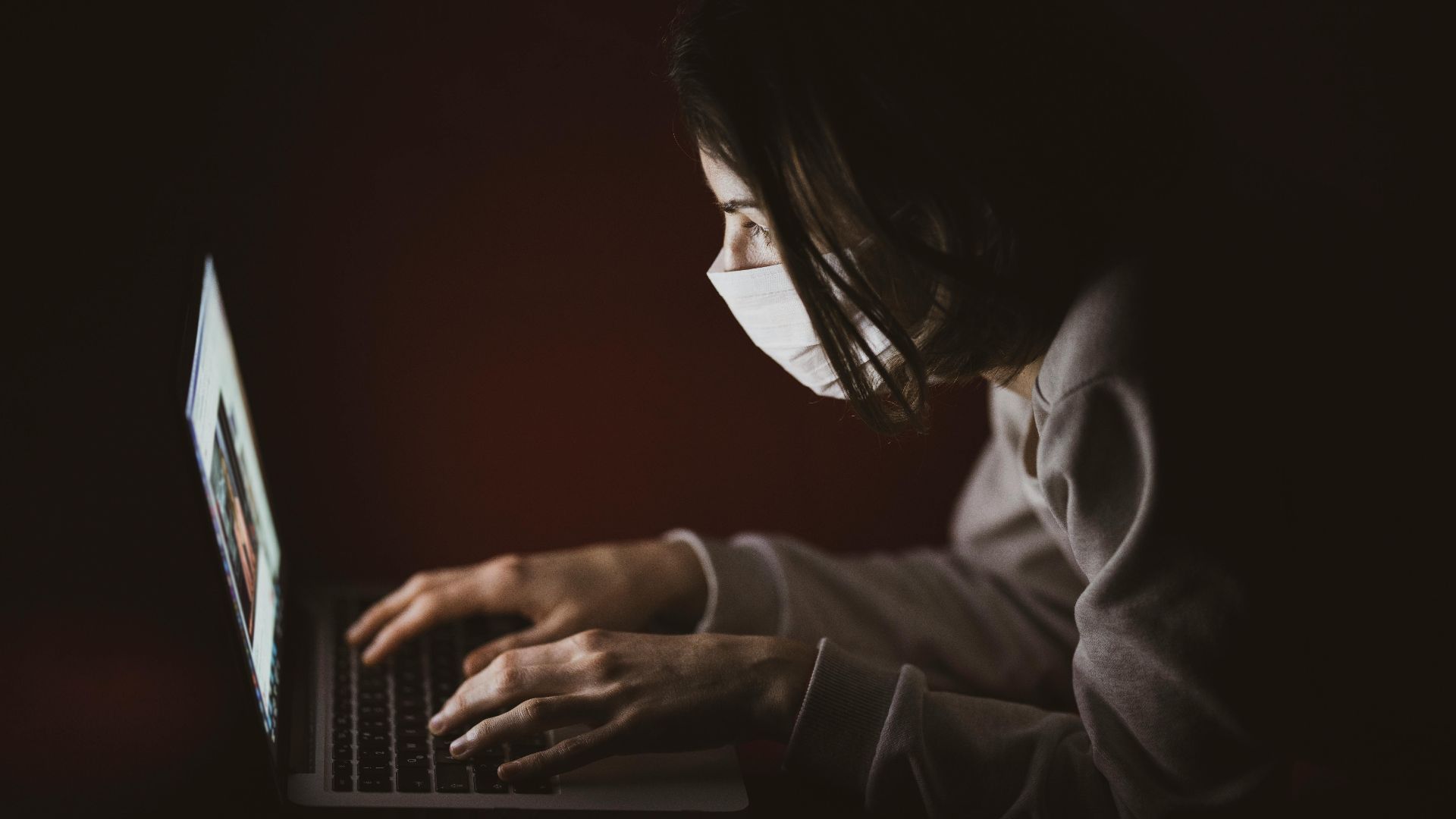 A woman wearing a face mask focuses on her laptop screen in dim lighting, highlighting remote work.