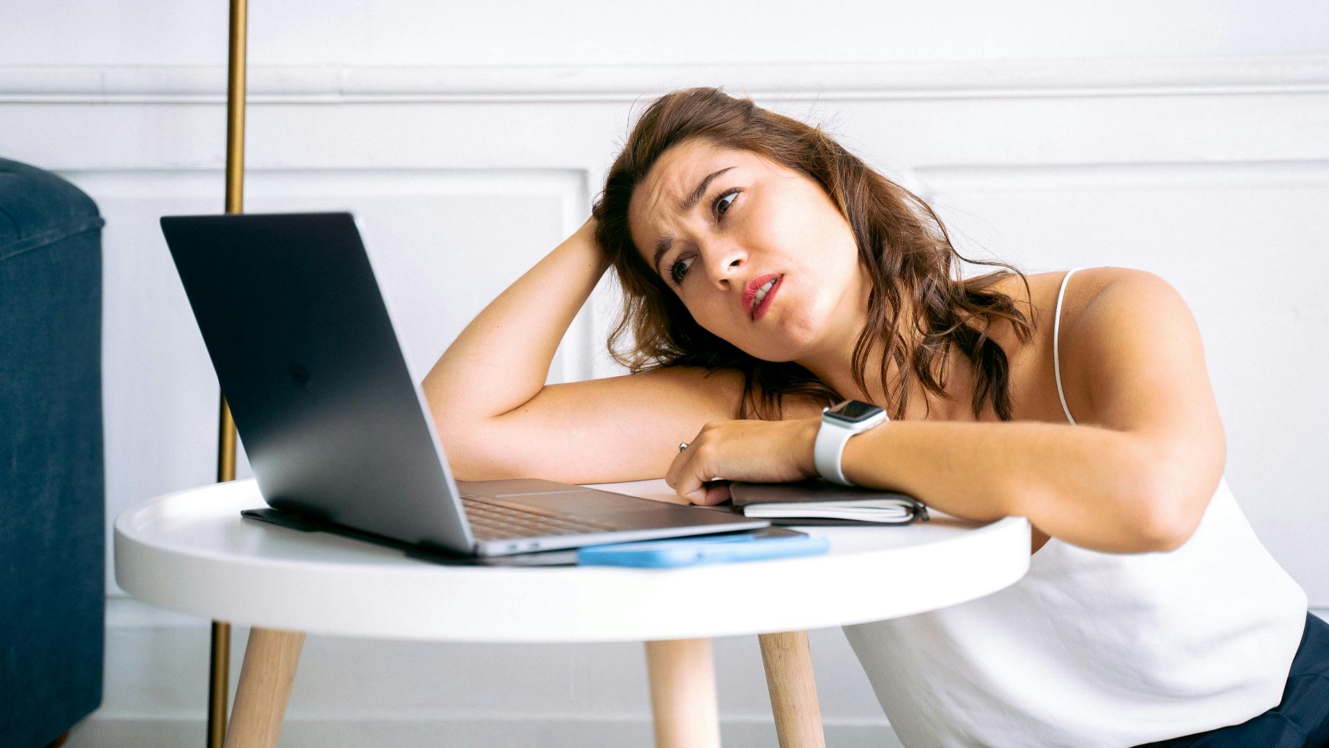 A frustrated woman sits at a desk with her hand on her head, staring at an open laptop.