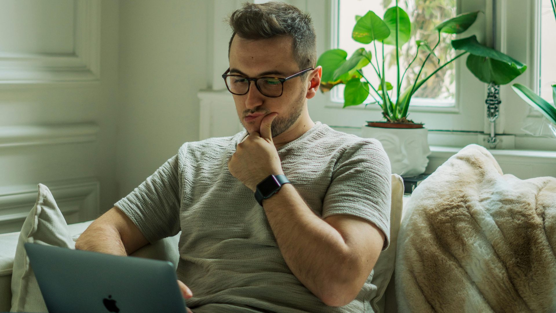 A man wearing eyeglasses using a laptop in a cozy home setting with natural light.