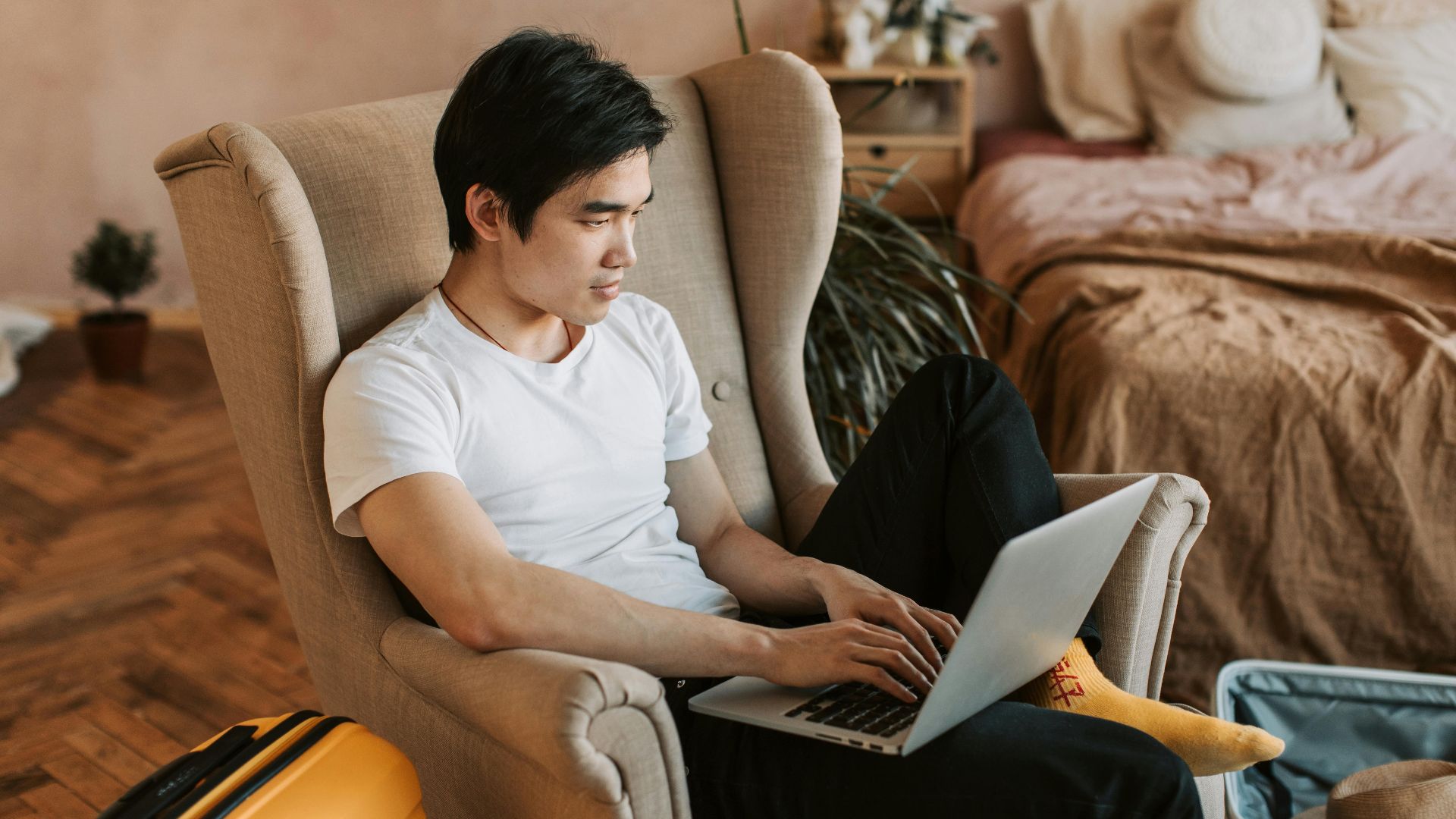 A man sitting comfortably in an armchair, using a laptop in a cozy home interior.