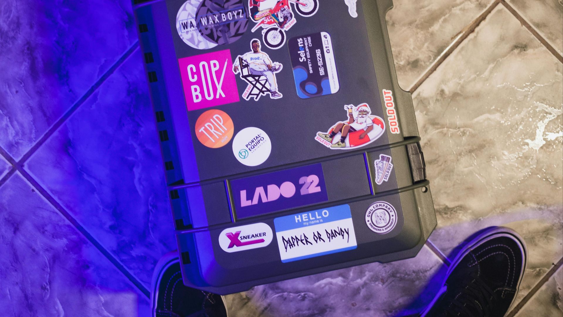 High-angle view of a suitcase covered in colorful stickers on a tiled floor next to a person's feet.