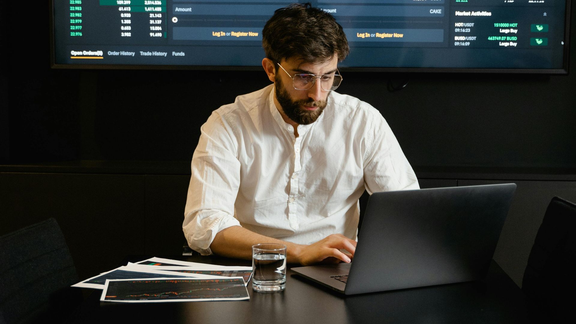 A focused man analyzing stock market trends on a laptop in an office setting with charts displayed.