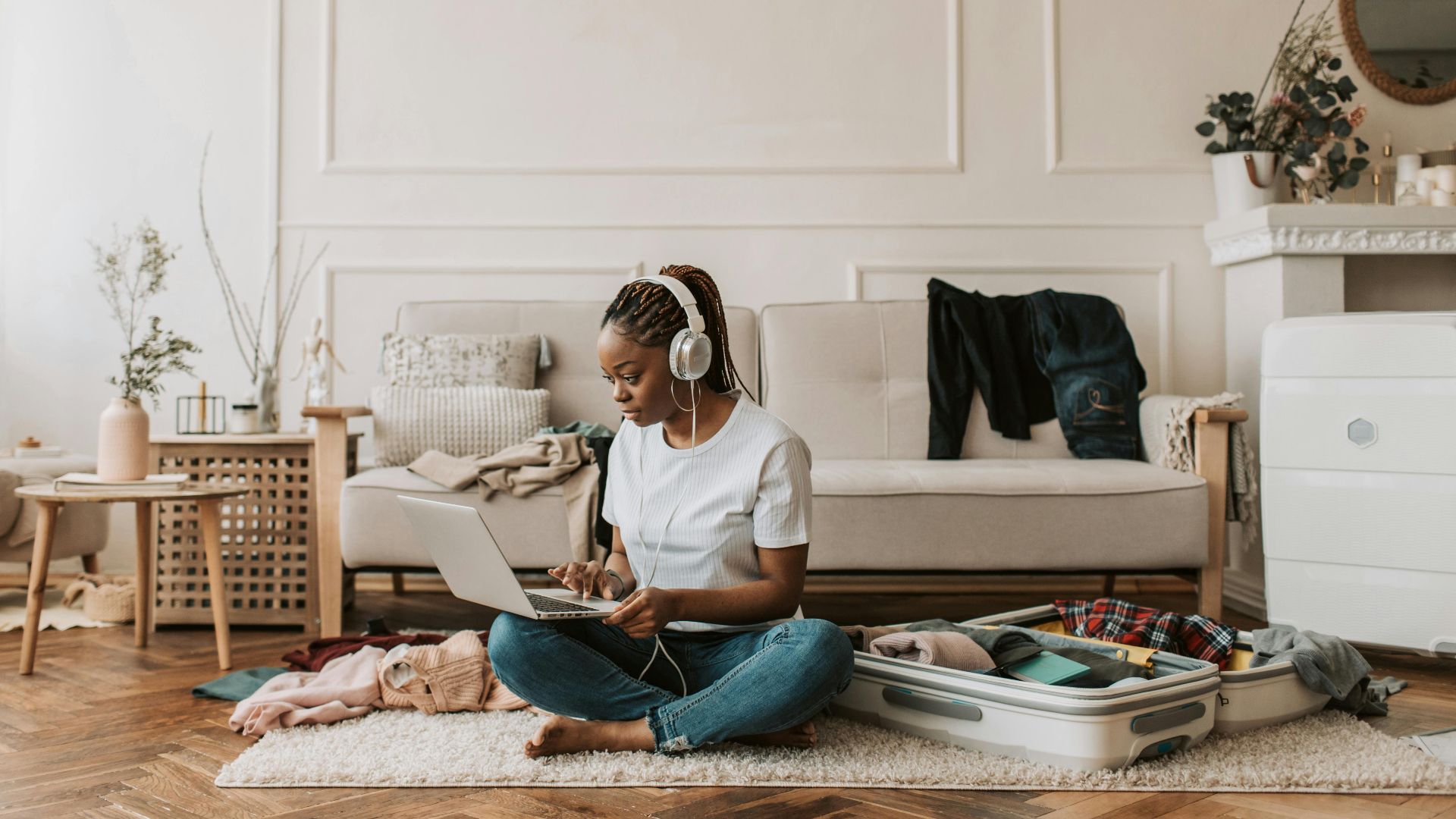 Young woman sitting on floor, packing suitcase, using laptop indoors.