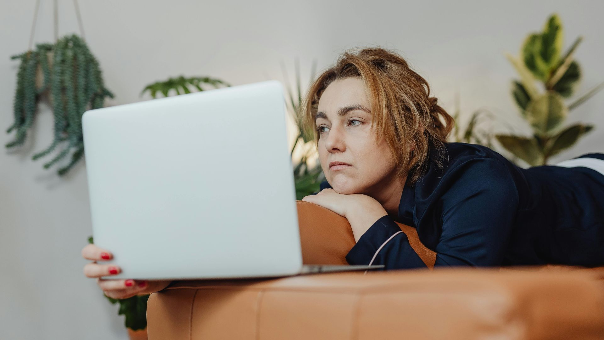 A woman lying on a couch indoors, looking upset while holding a laptop in a cozy home environment.