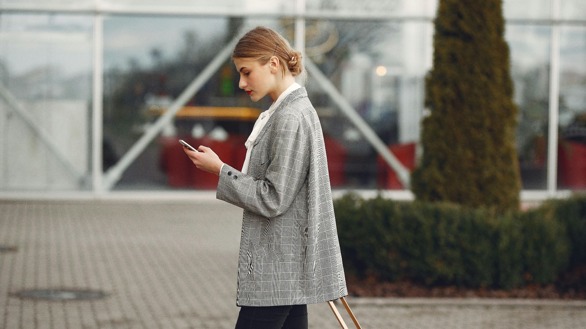 Stylish woman walking with suitcase while using smartphone outside a modern building.