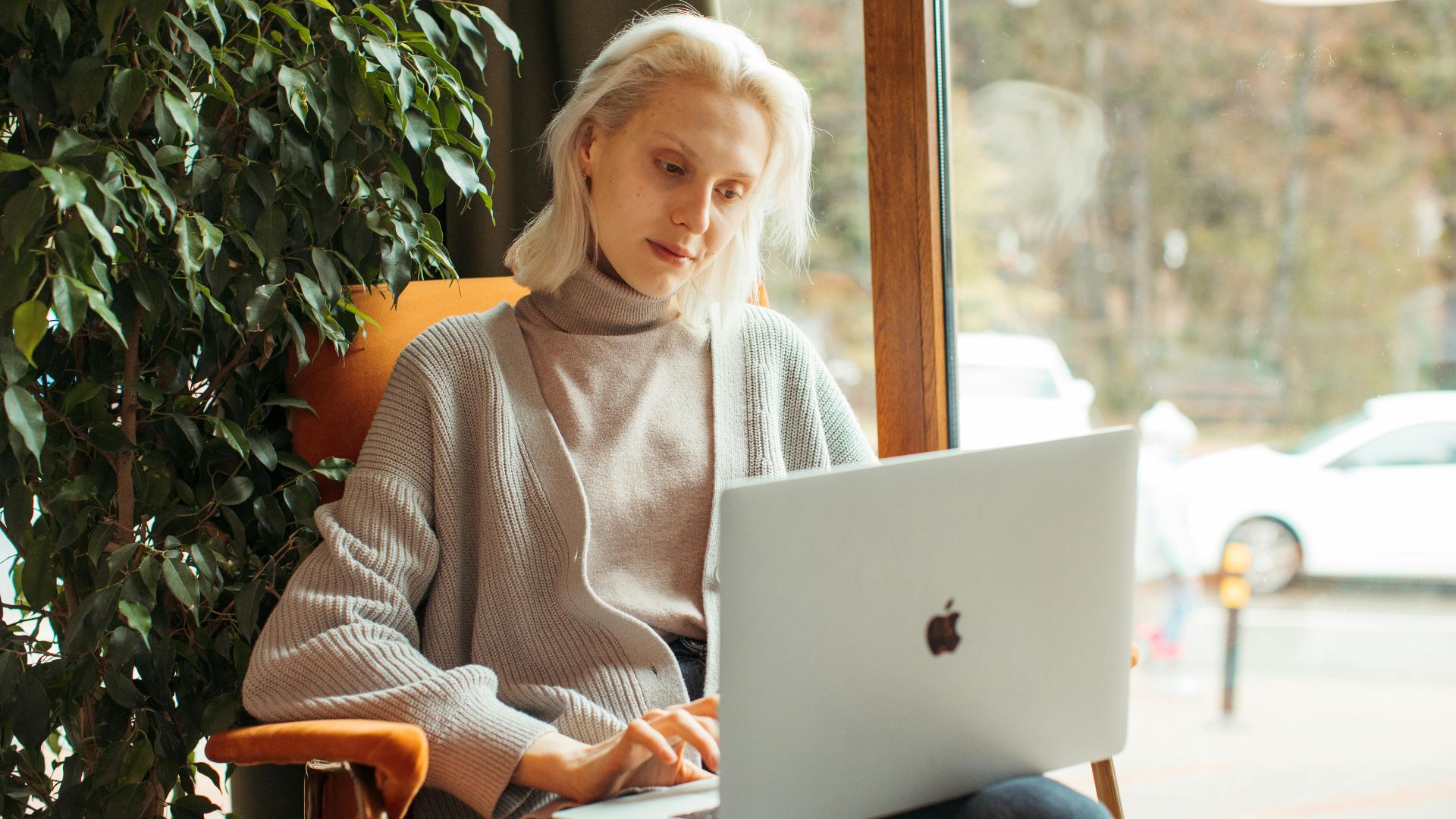 Blonde woman working on a laptop in a cozy office setting with indoor plants.