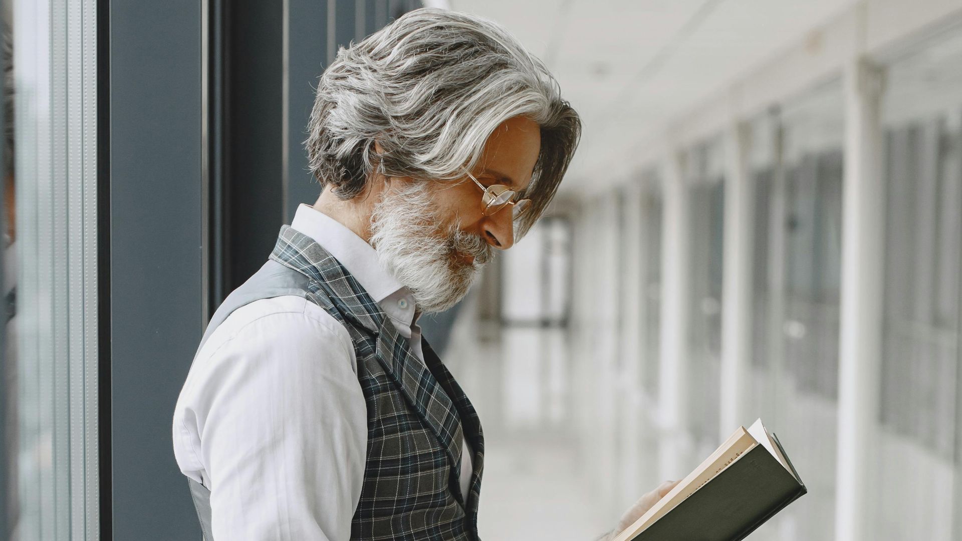 A mature man with gray hair reading a book in an airport hallway, leaning on luggage.