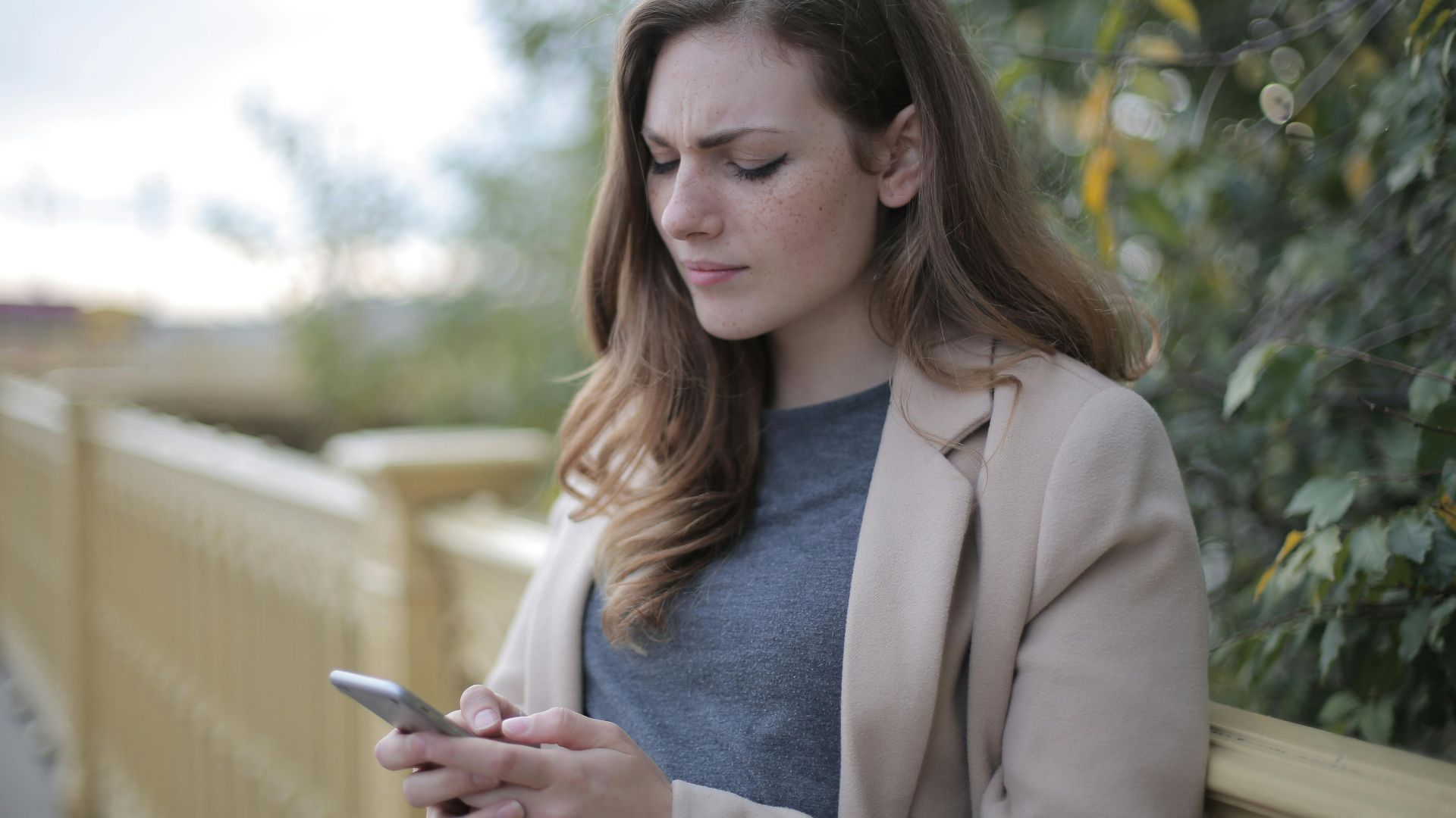 A woman stands outdoors in a casual outfit, using her smartphone with a pensive look.