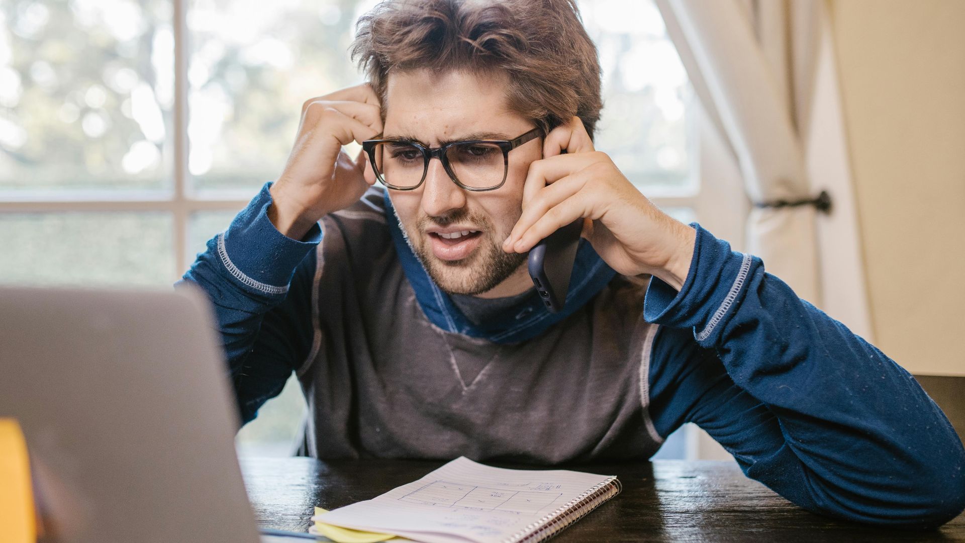 Man in glasses using laptop and smartphone at home, appearing confused.