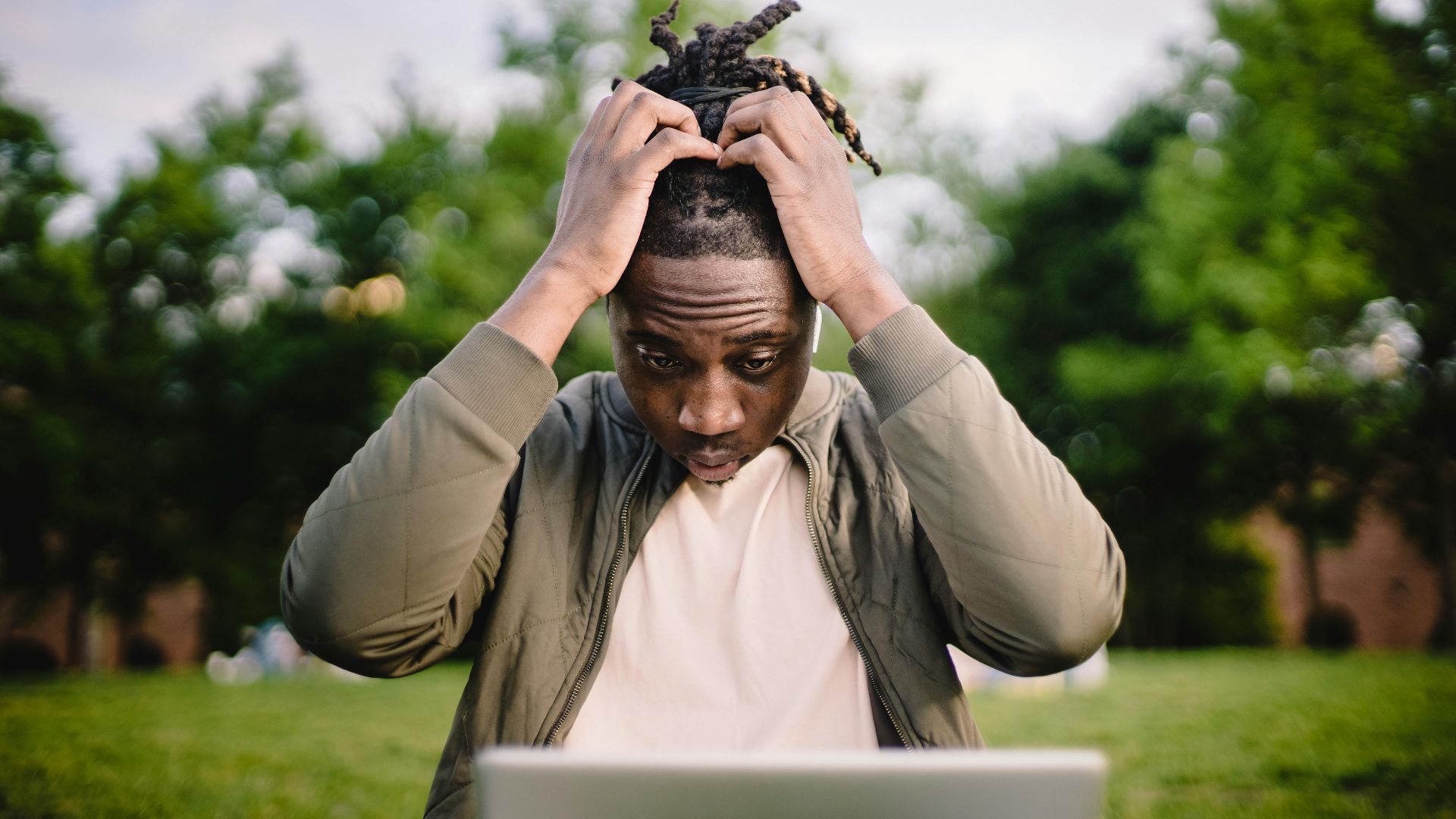 Upset young African American male freelancer clutching head with hands after failure in project while working remotely in park