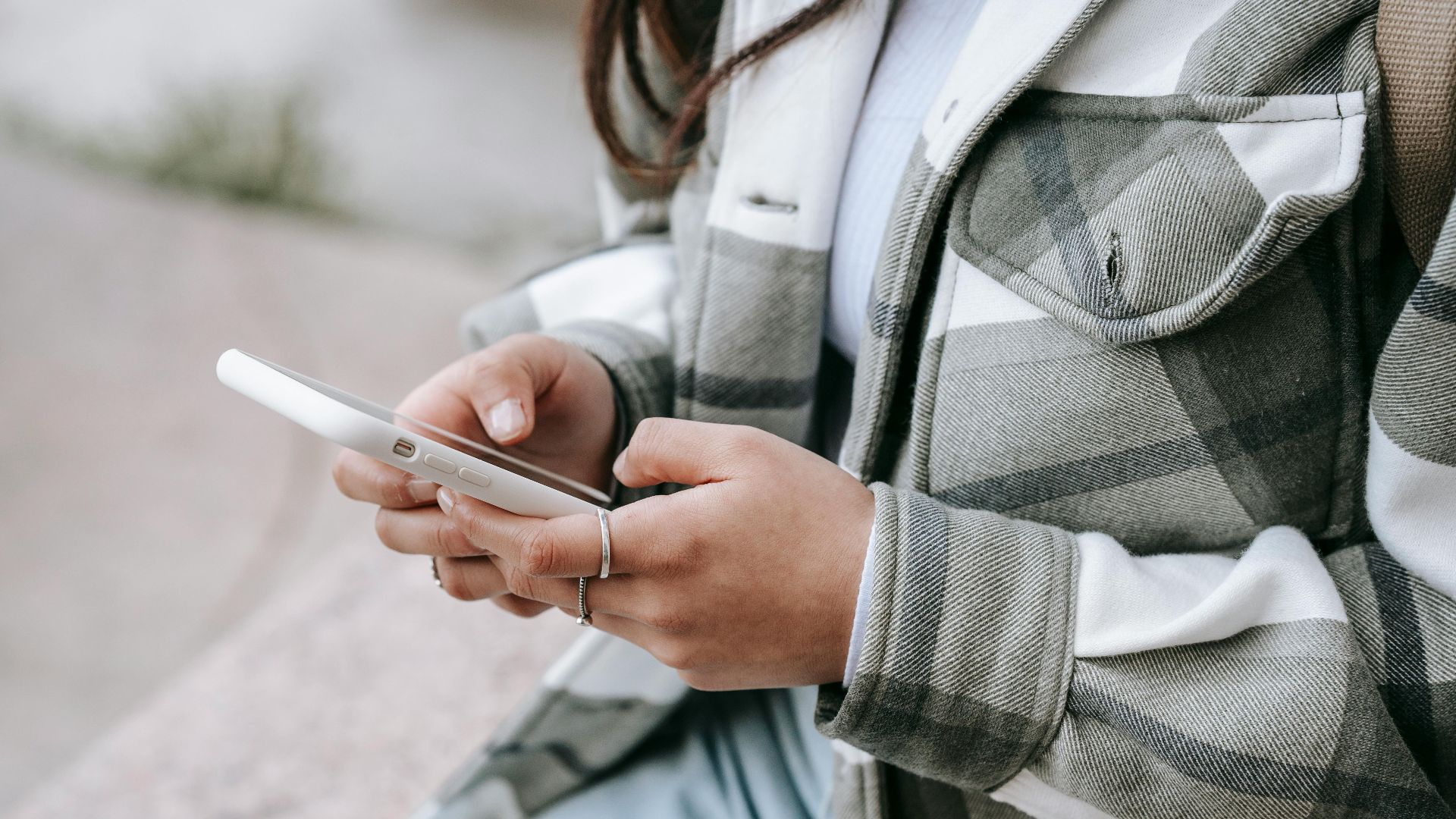 Crop anonymous young woman in casual outfit sitting on stone fence and text messaging on phone in daytime