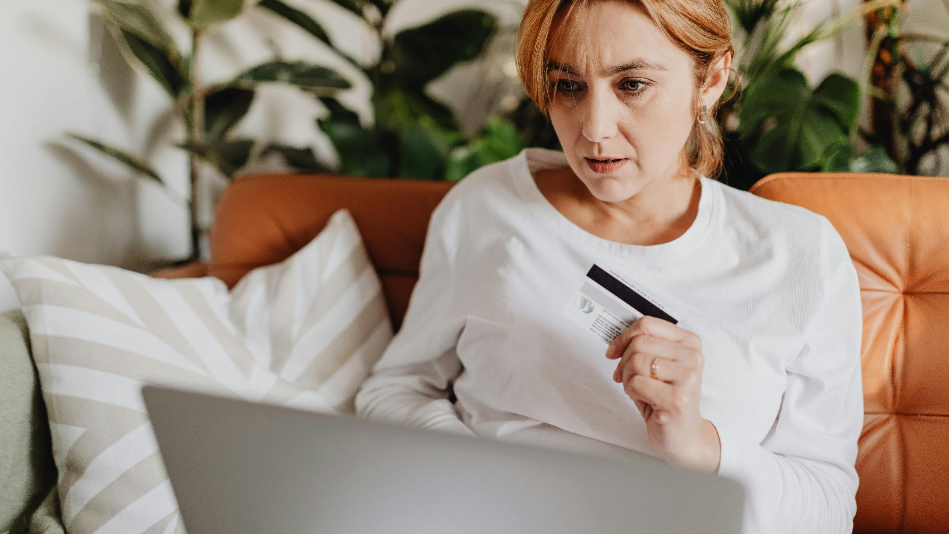 A woman sitting on a couch shopping online with a credit card and laptop in a cozy living room.