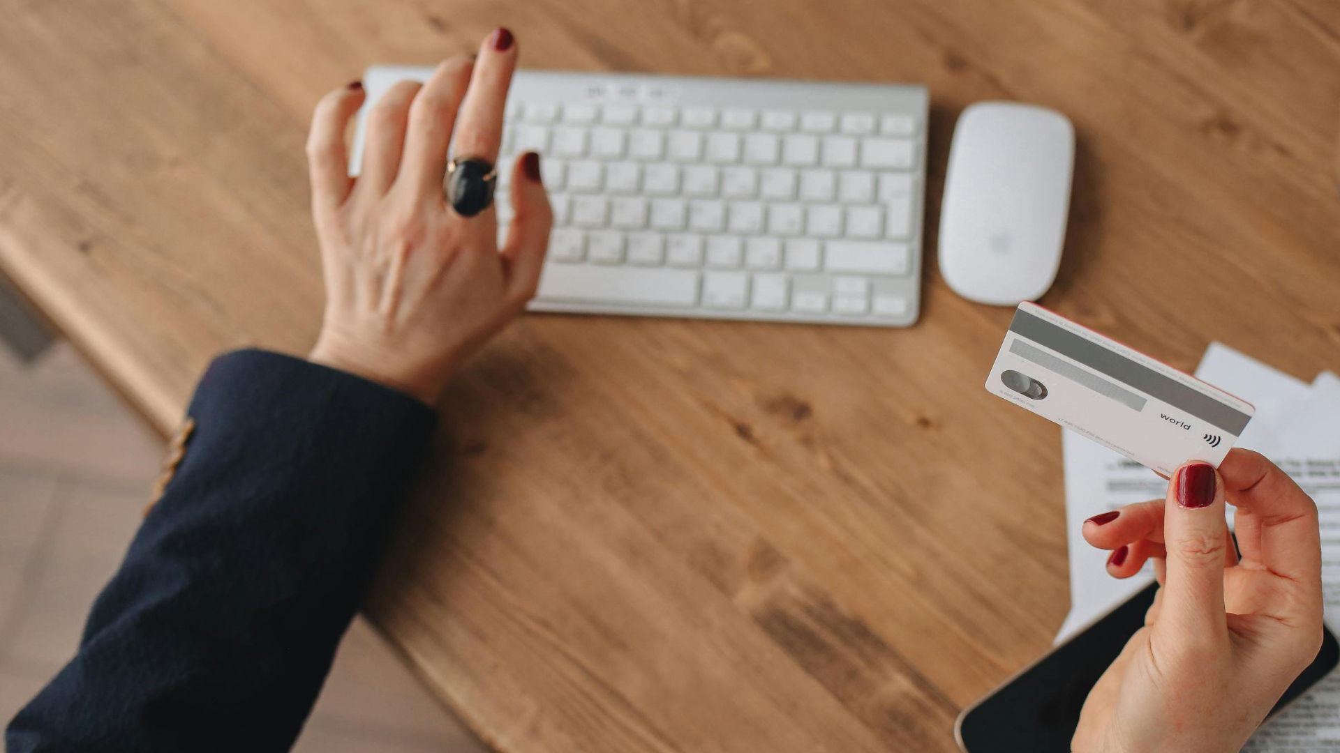 A woman uses an Apple Magic Keyboard while holding a credit card over paperwork on a wooden desk.