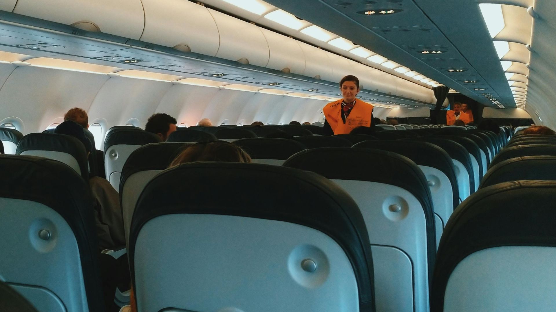 Inside of modern airplane cabin with passengers sitting on comfortable seats and cabin crew standing at passageway