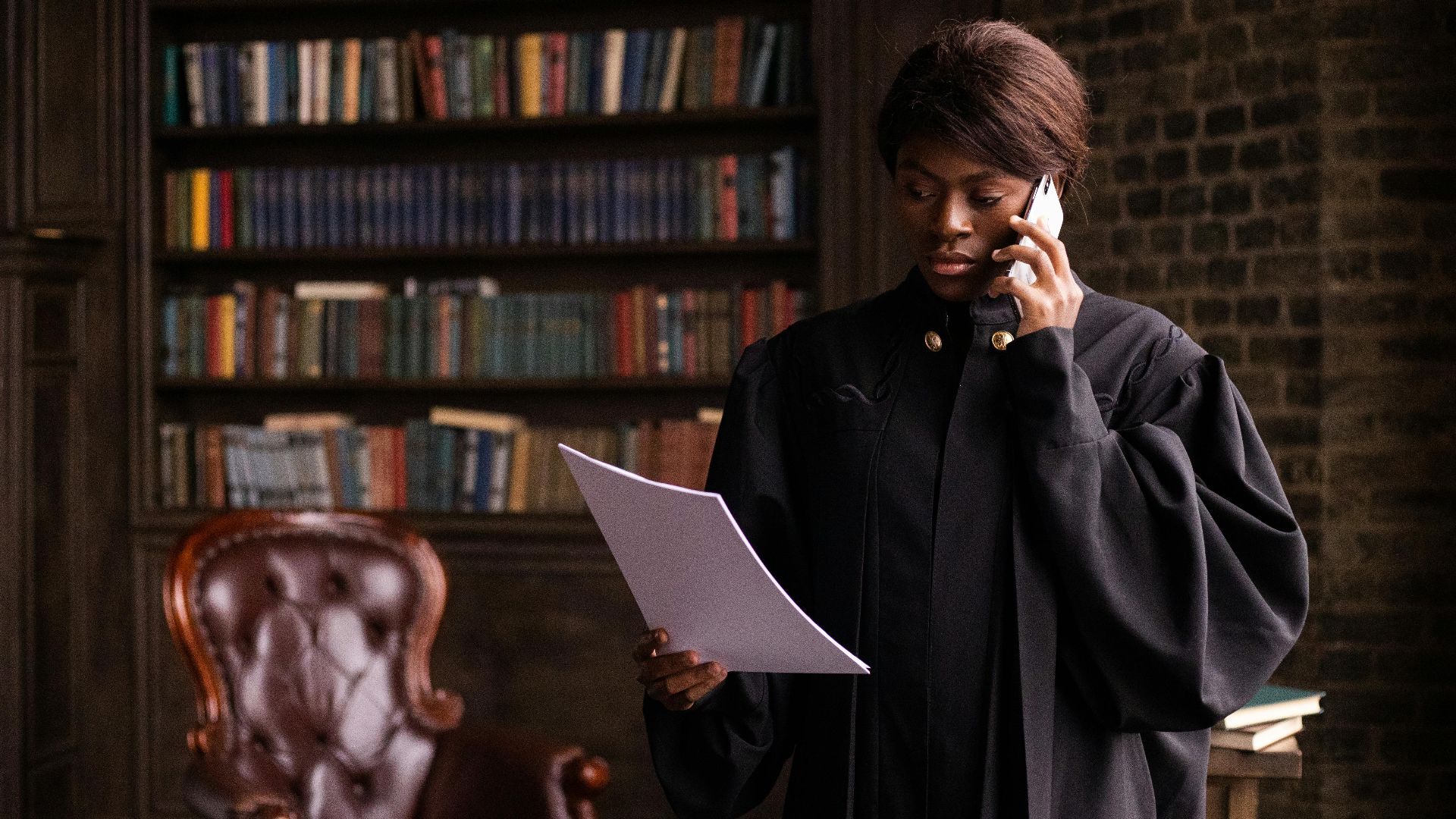 African American judge in traditional robes holds a document and talks on the phone in a law library.