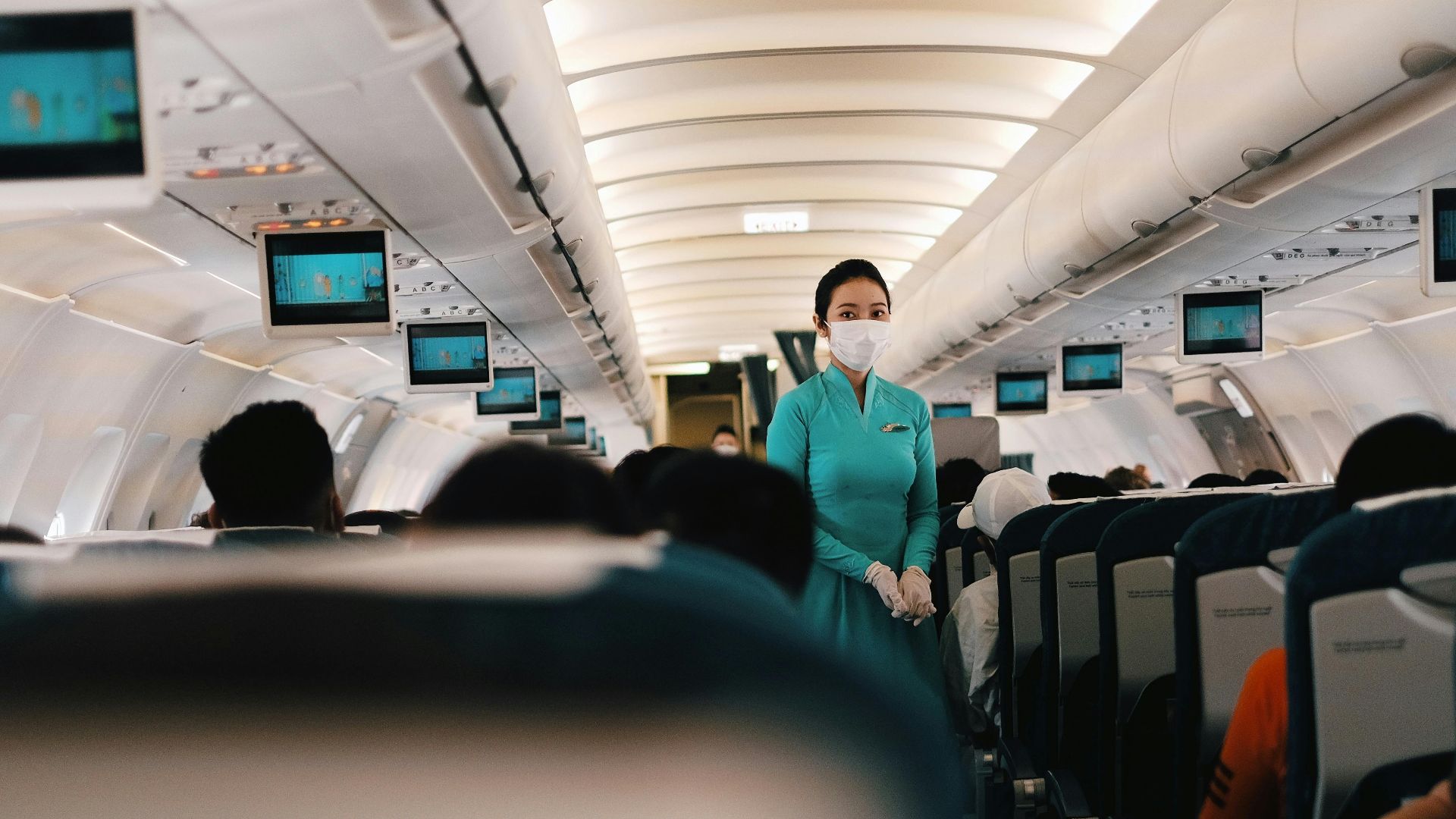 Flight attendant wearing a face mask attending to passengers in an airplane cabin.