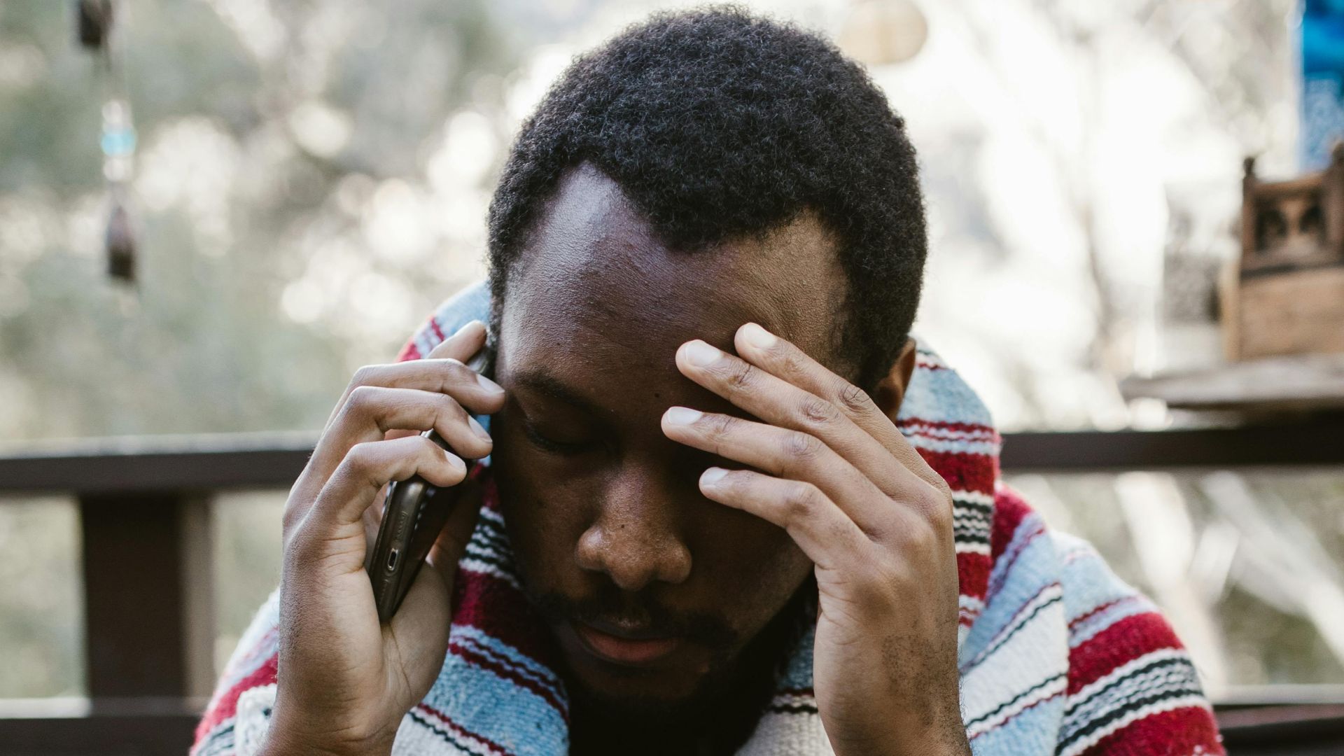 A man wrapped in a blanket talks on the phone, looking stressed in an outdoor setting.