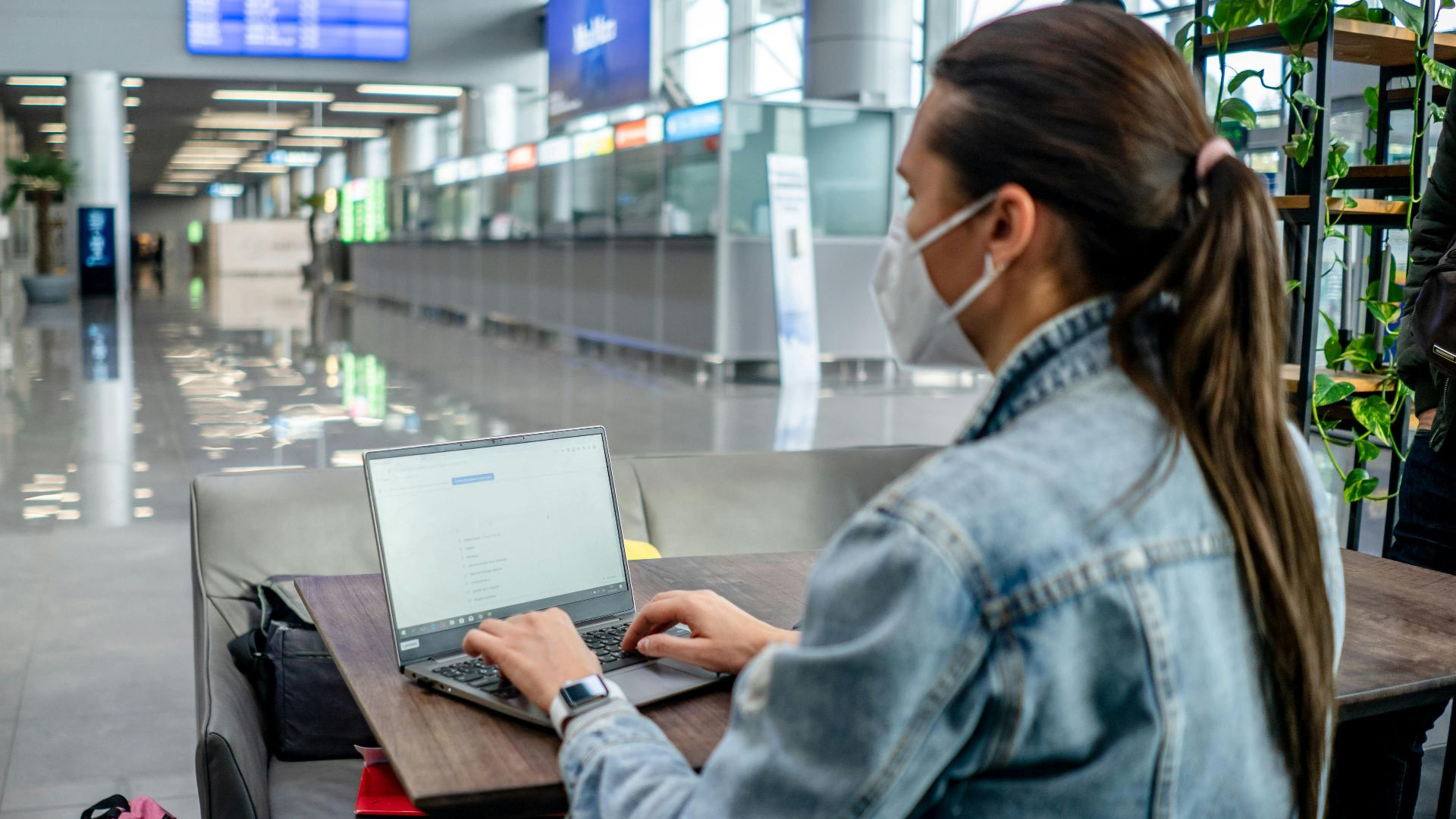 Side view of concentrated female freelancer in protective mask typing on netbook while sitting at table in modern airport terminal