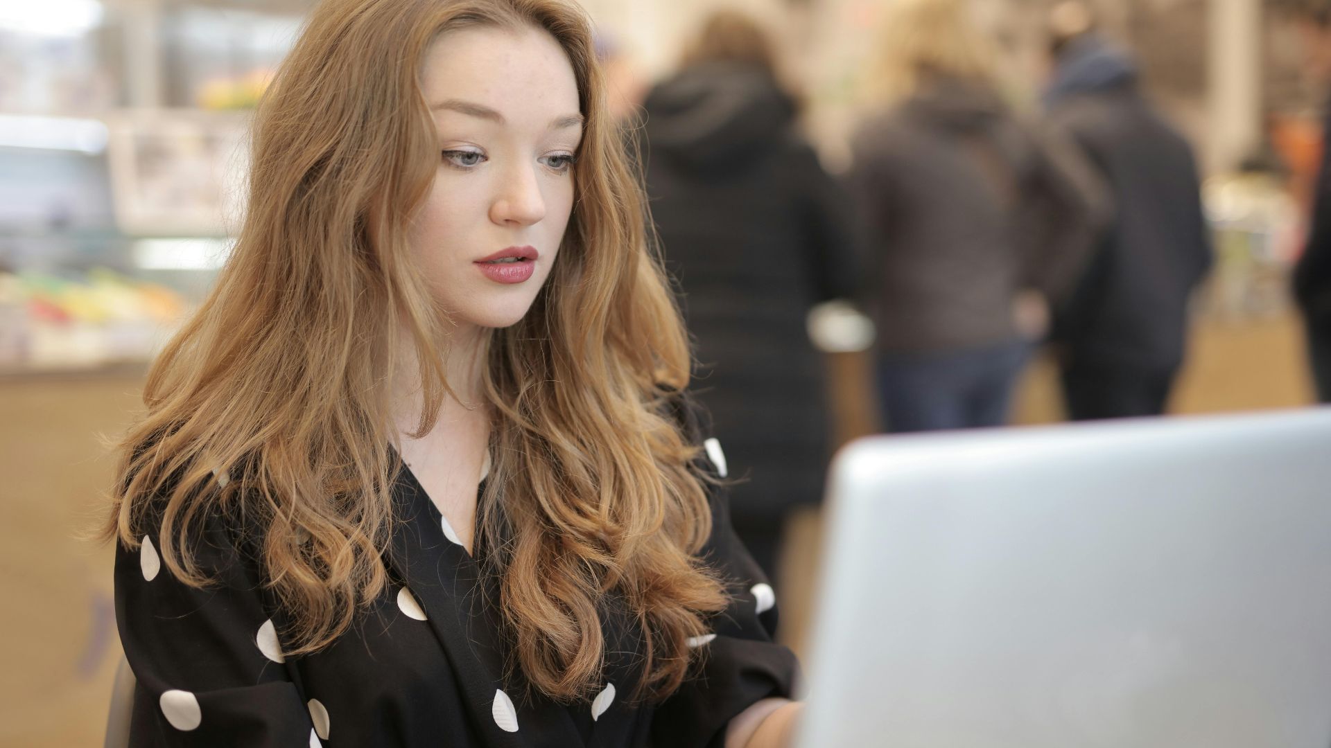 A focused woman in a polka dot shirt is typing on a laptop in a bustling cafe.
