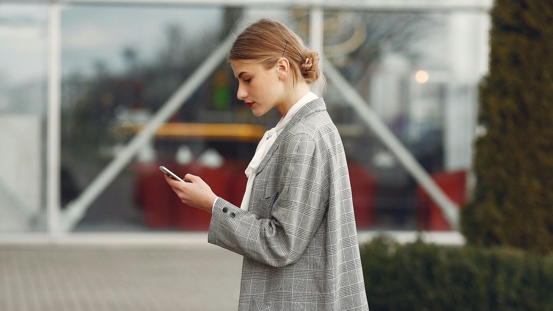 Stylish woman walking with suitcase while using smartphone outside a modern building.