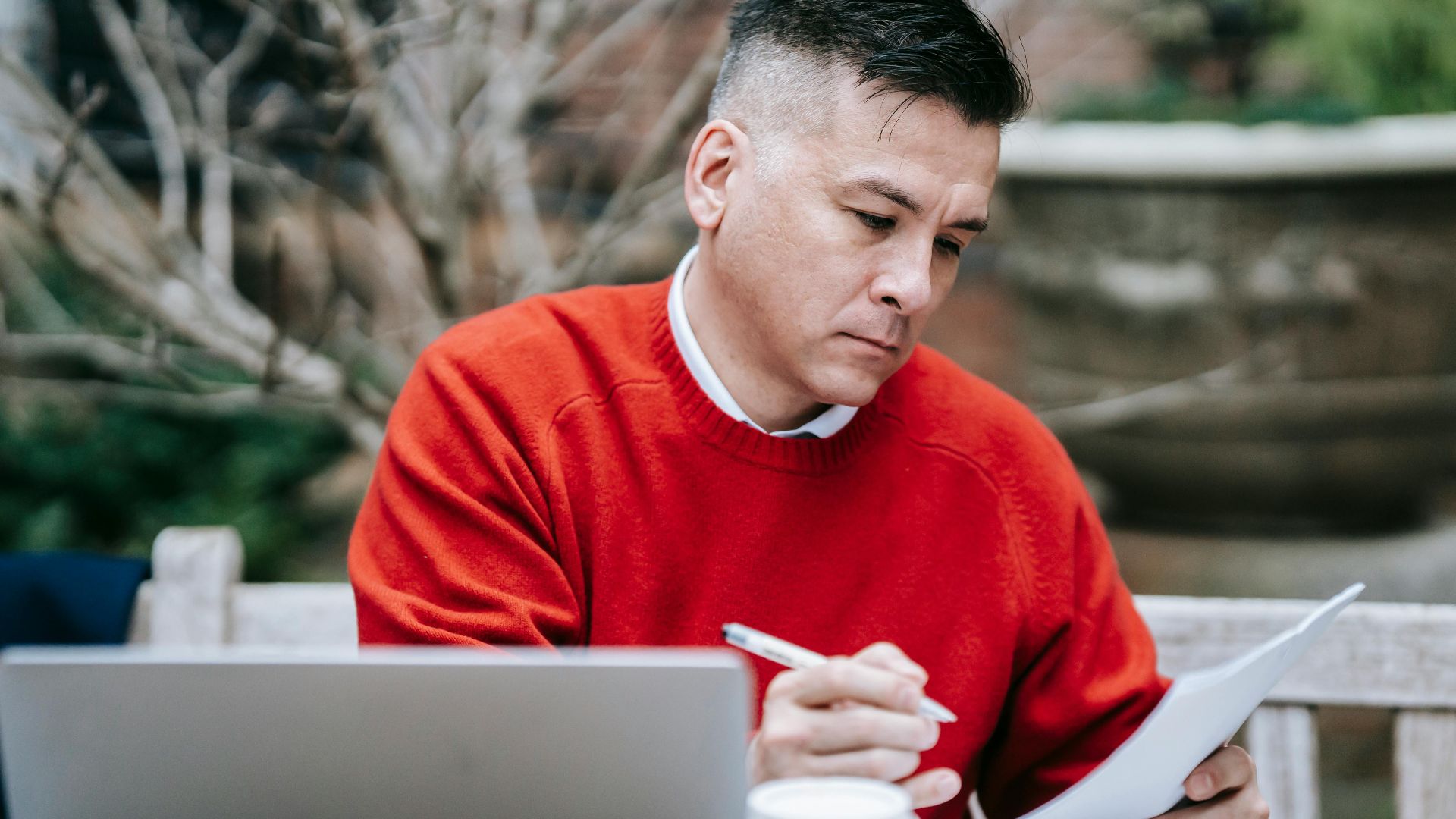 A man in a red sweater concentrating on paperwork while working remotely with a laptop outdoors.