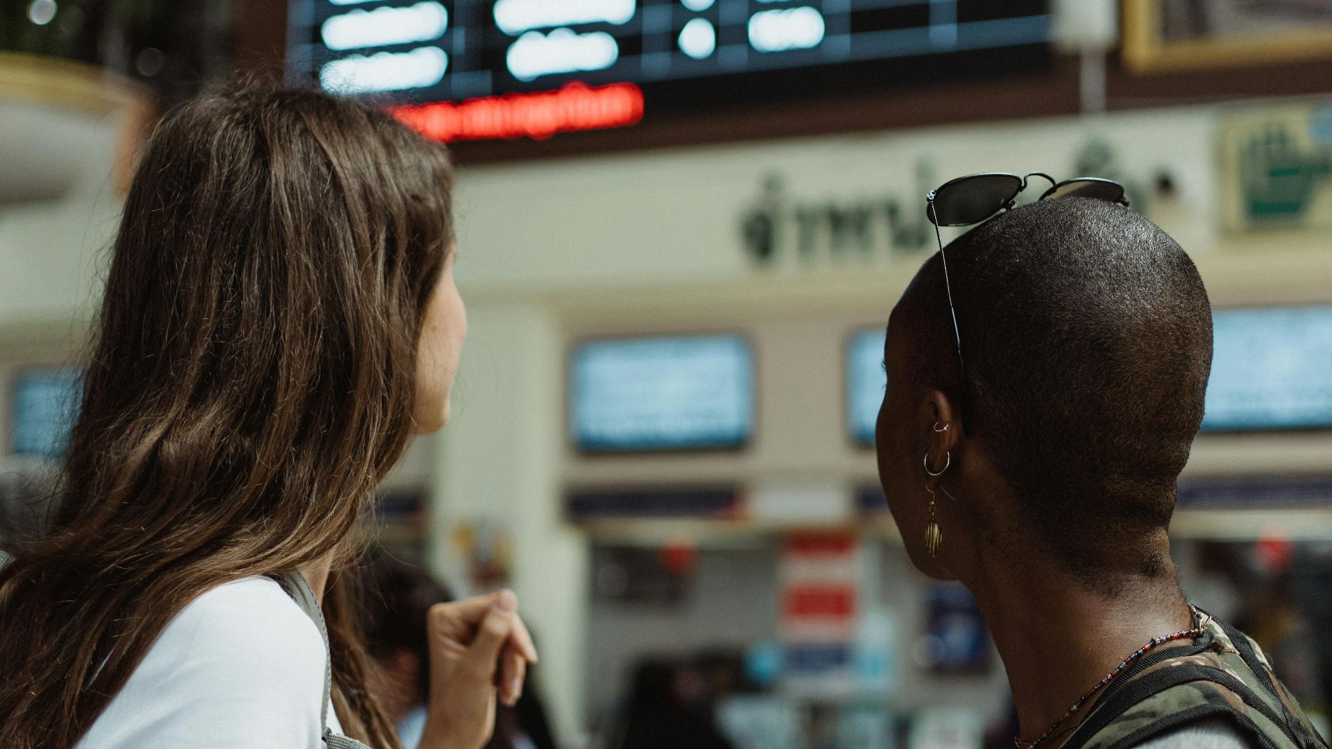 Two women checking the flight schedule on a departure board in an airport terminal, preparing for travel.
